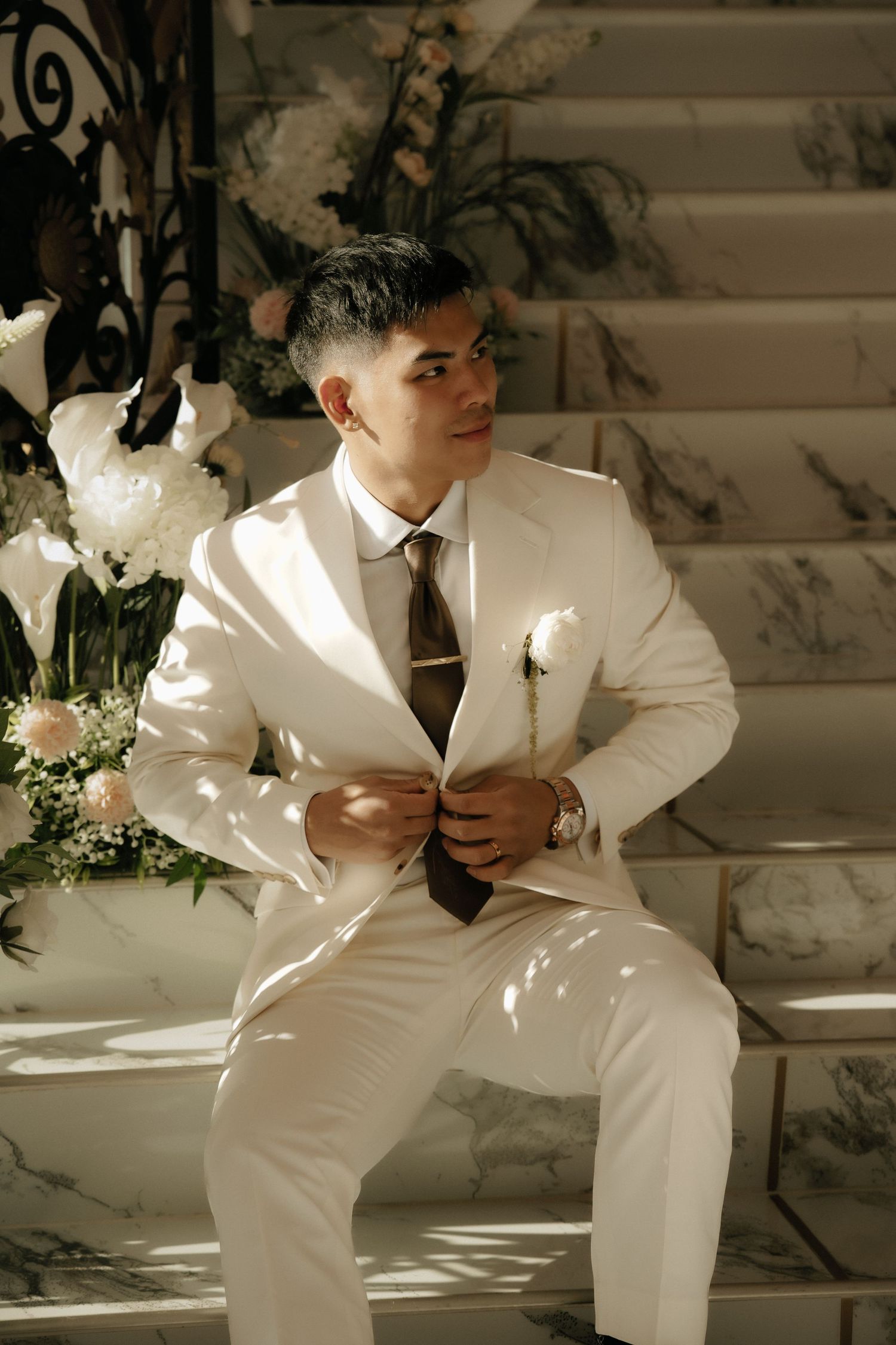 A young man in a white suit with a floral boutonniere sits on marble steps surrounded by white flowers.