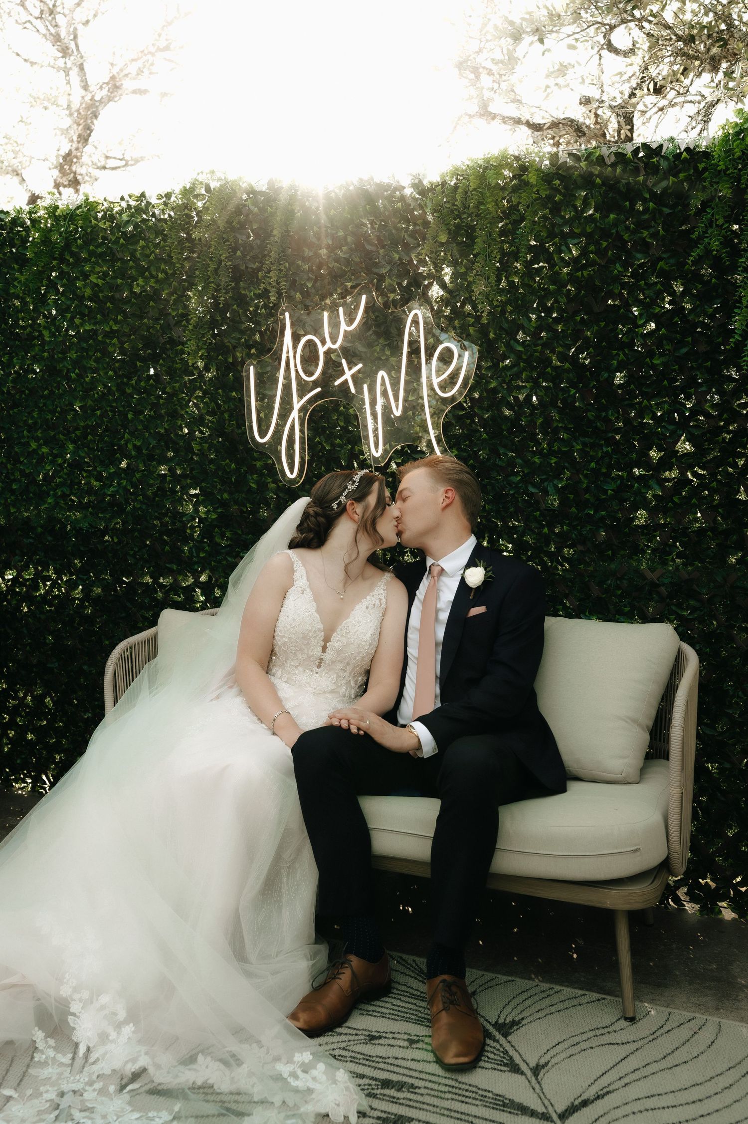 Couple shares romantic moment on white loveseat against garden hedge backdrop.