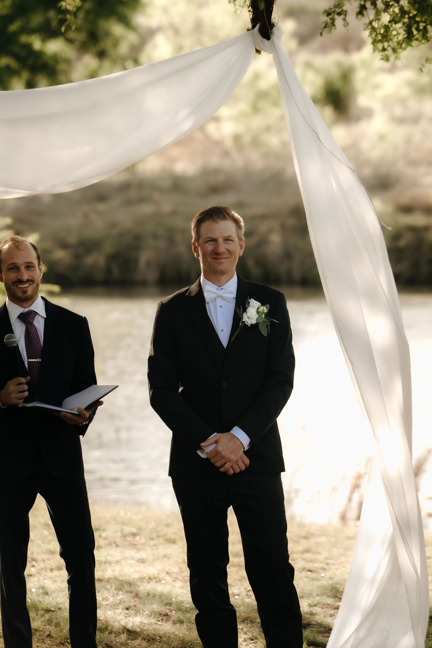 Groomsmen in black suits standing in a line outdoors with flowing white fabric decor.