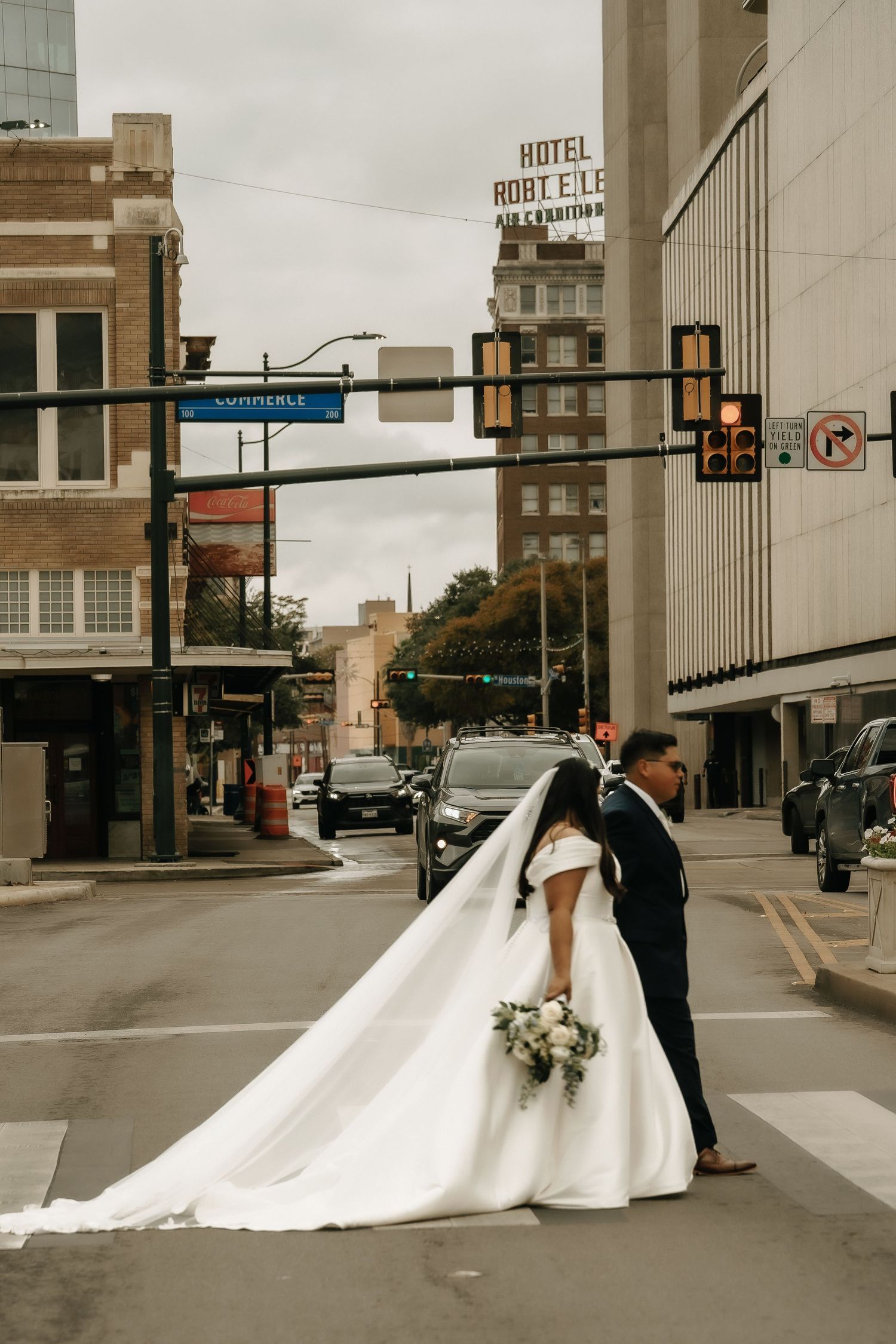 A couple in wedding attire walks together on a city street with tall buildings and a hotel sign in the background.