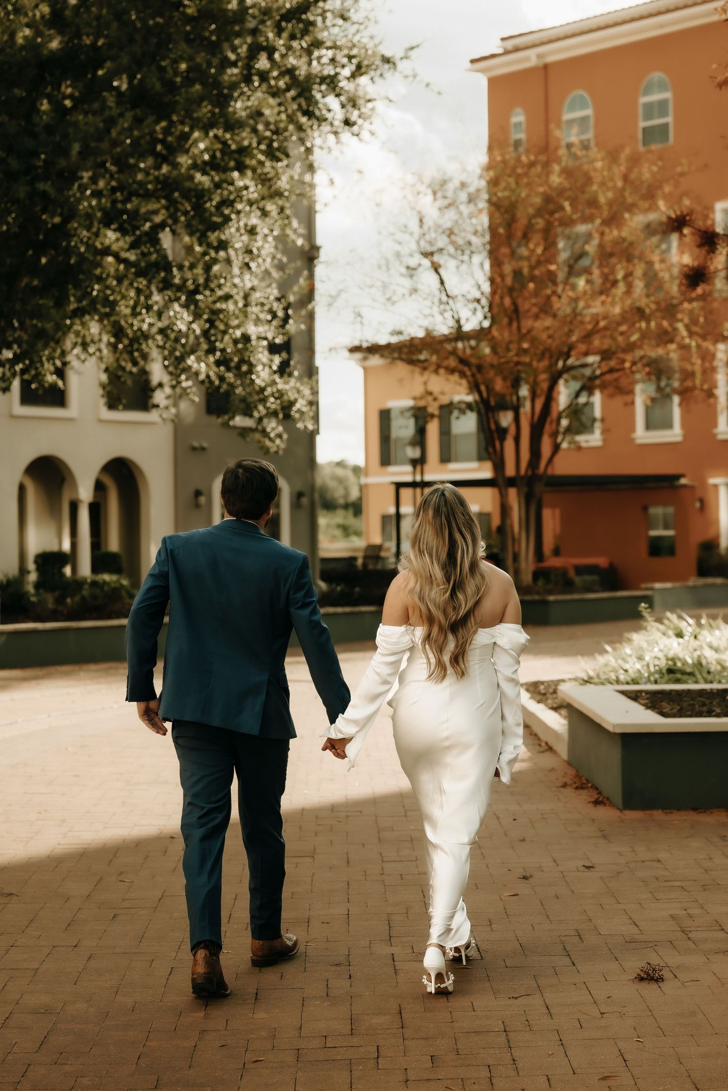Couple walking hand in hand on brick pavers with orange buildings in background.