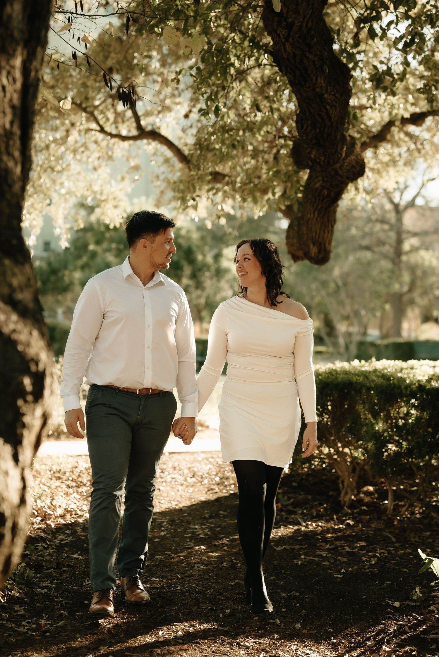 Couple walking together through sunlit garden path in casual attire.
