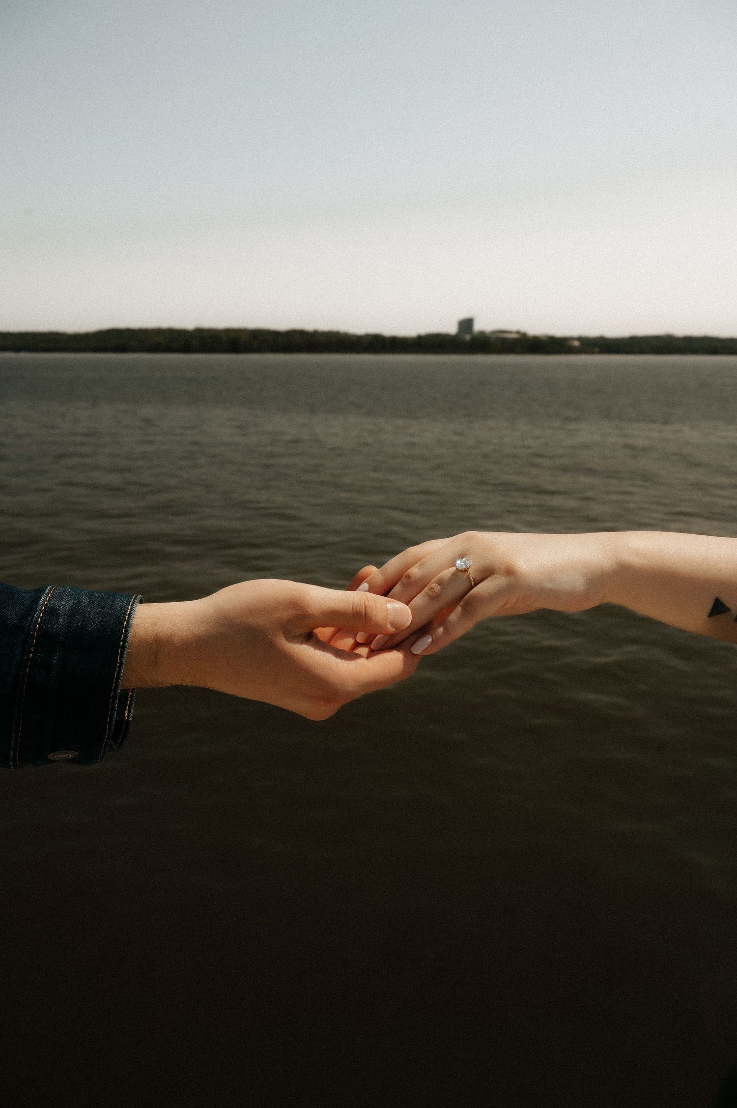 Two hands gently touching over water, woman wearing an engagement ring, romantic proposal moment outdoors.