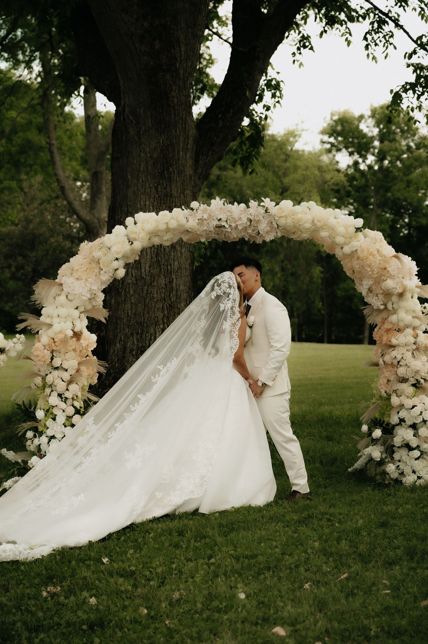 Bride and groom kissing under a floral arch with white blooms outdoors near a large tree on their wedding day.