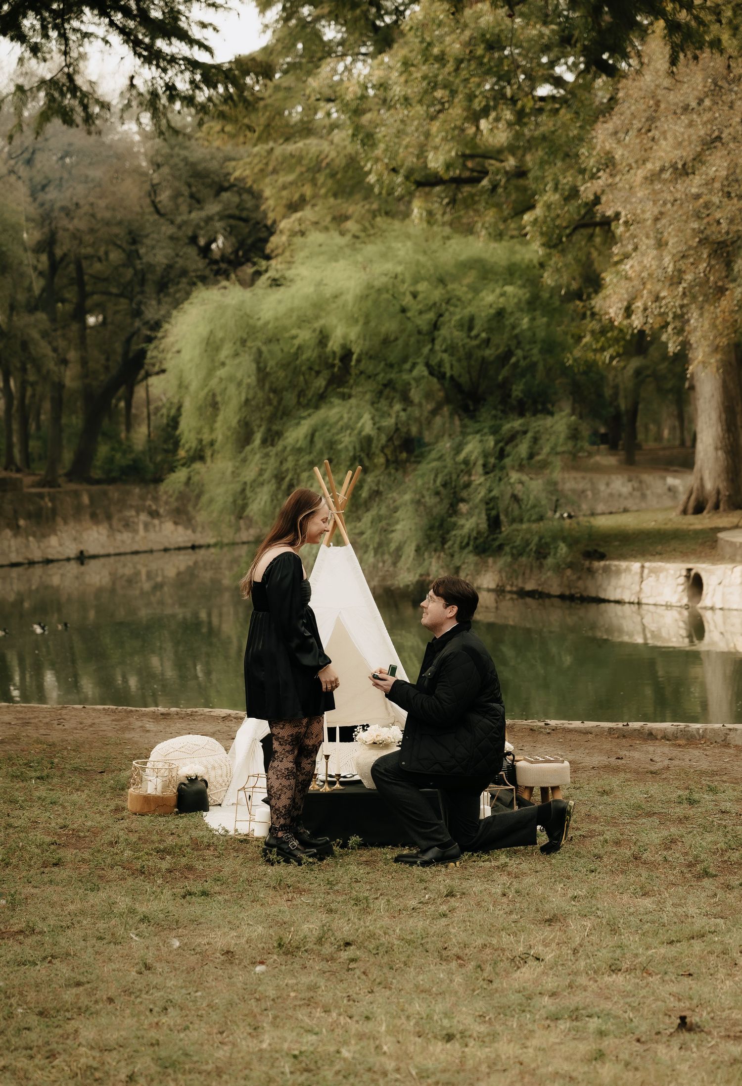 A couple shares a romantic picnic proposal by a lake in an autumn park setting.