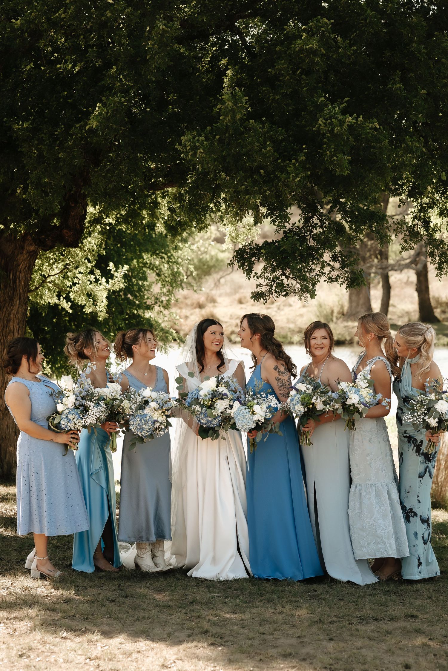 Bridal party in coordinating blue and white dresses pose under trees with bouquets.