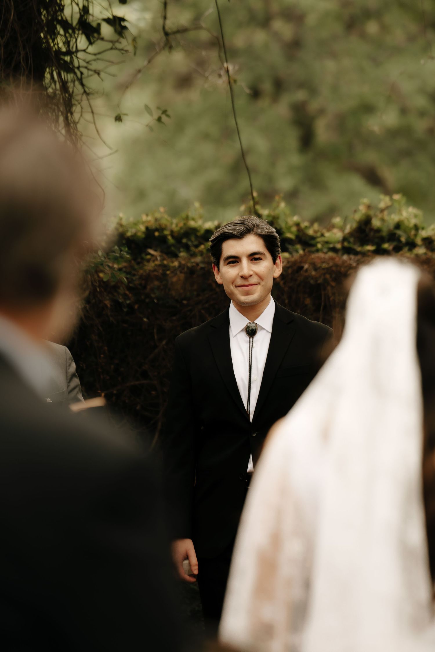 Wedding ceremony guests gather outdoors with greenery in the background.