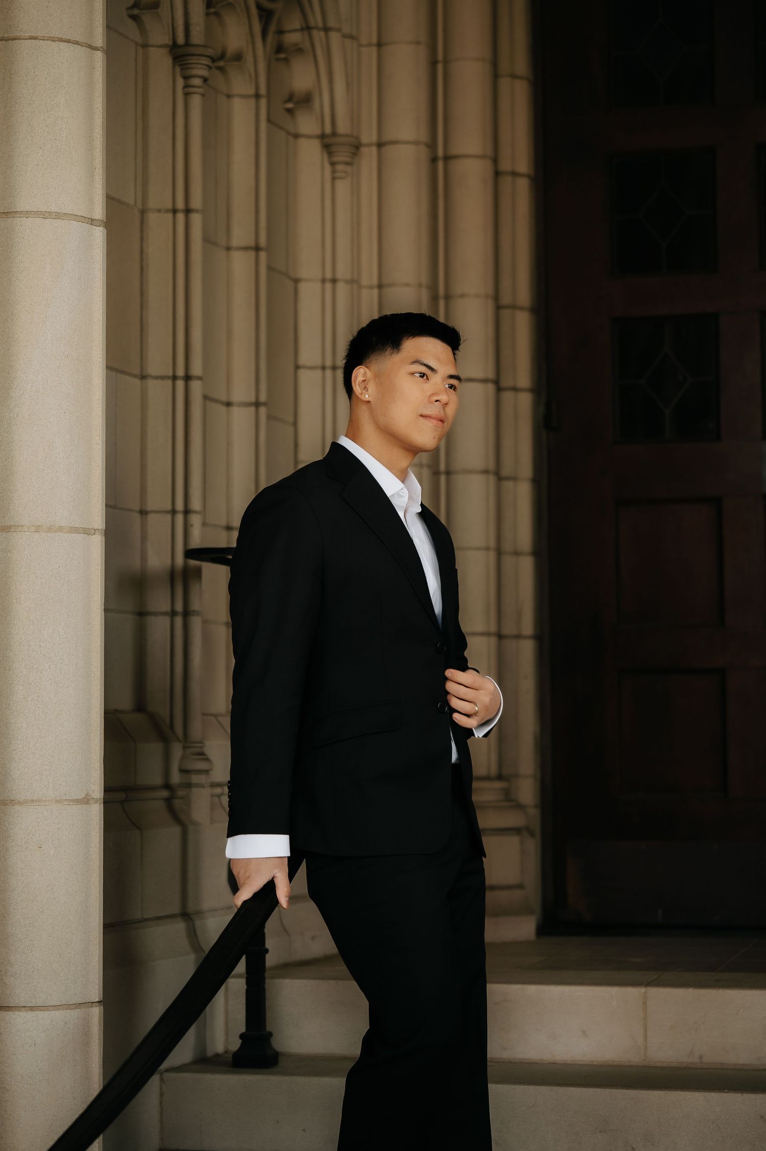 Dapper groom in black tuxedo stands confidently on steps of a grand stone building.