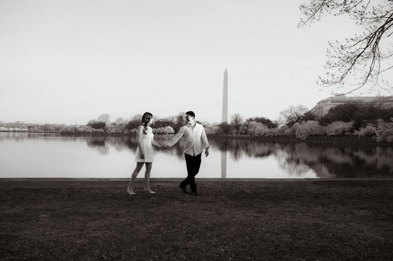 Couple holds hands walking by the Tidal Basin with Washington Monument in black and white photo.