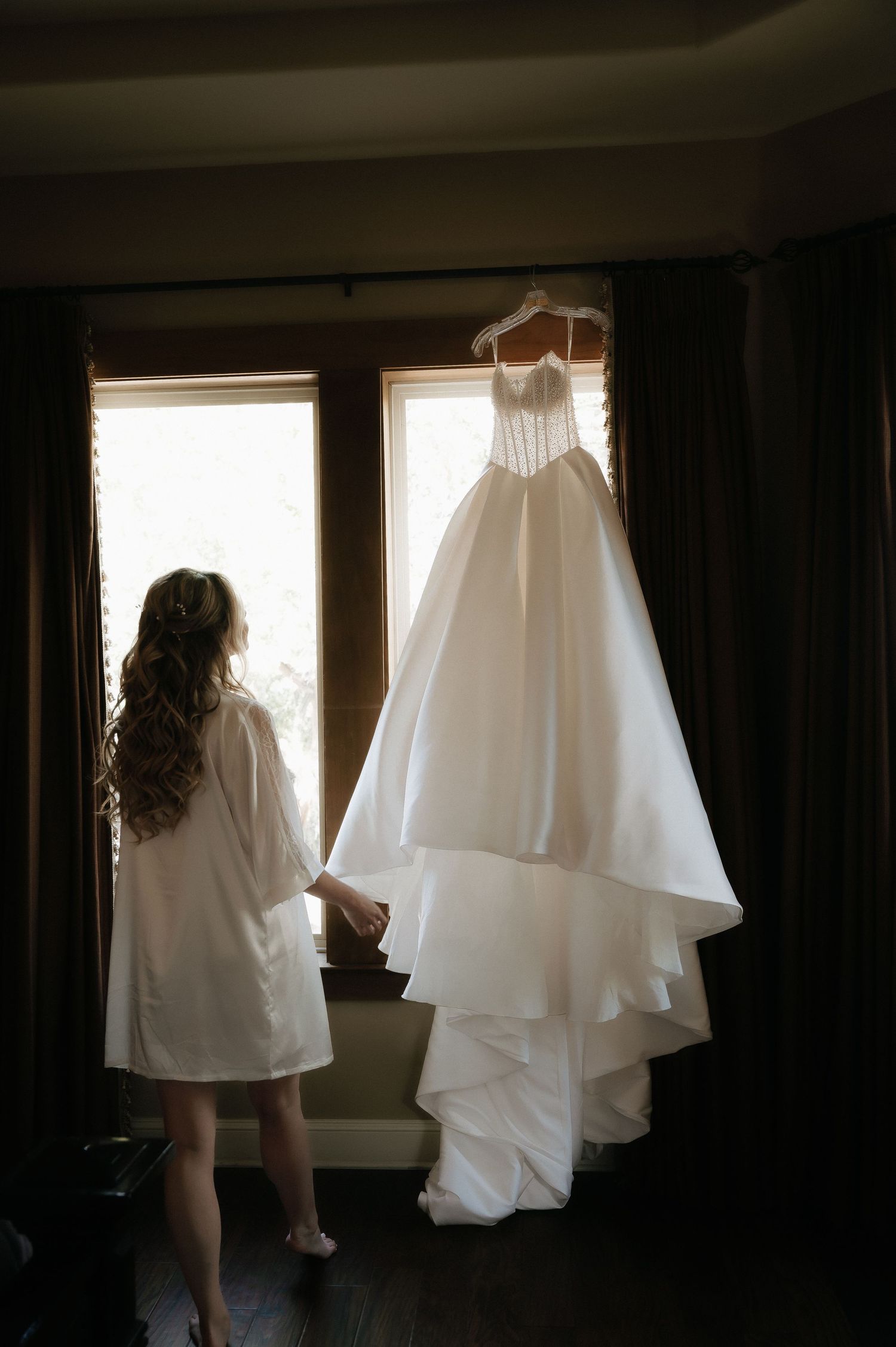 White wedding dress hanging by window with dramatic backlighting and dark curtains.
