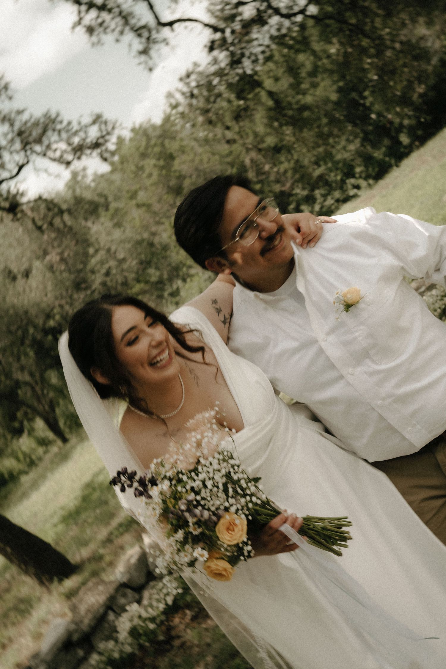 A joyful wedding couple laughing together outdoors while holding a bouquet of baby's breath flowers.
