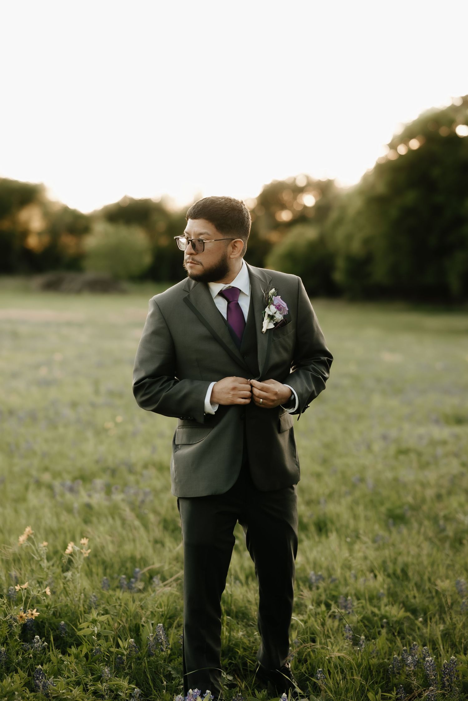 Groomsmen in dark suits share laughs while standing in a sunlit field.