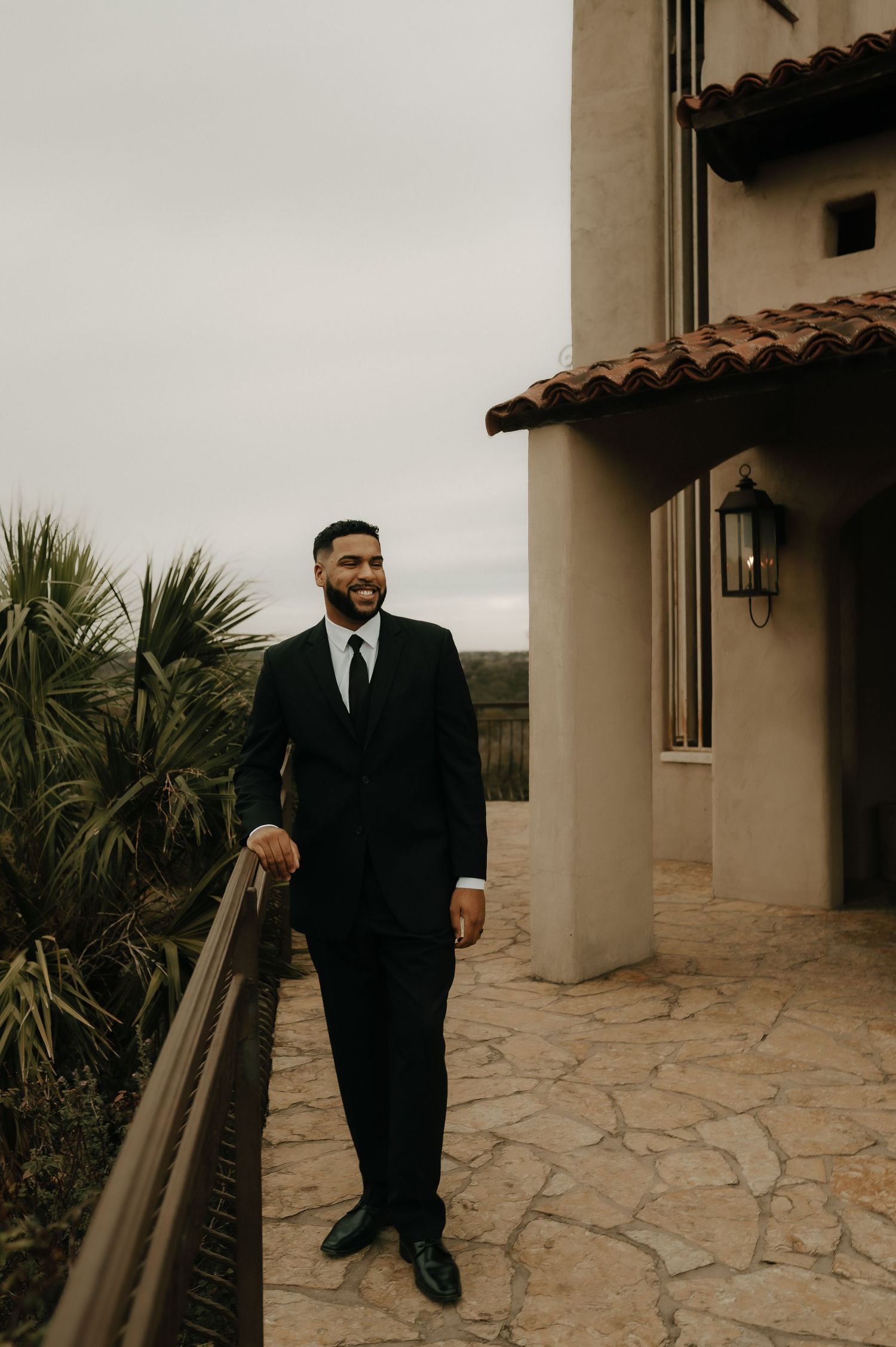 Groom in black suit standing on stone patio of Mediterranean-style building.