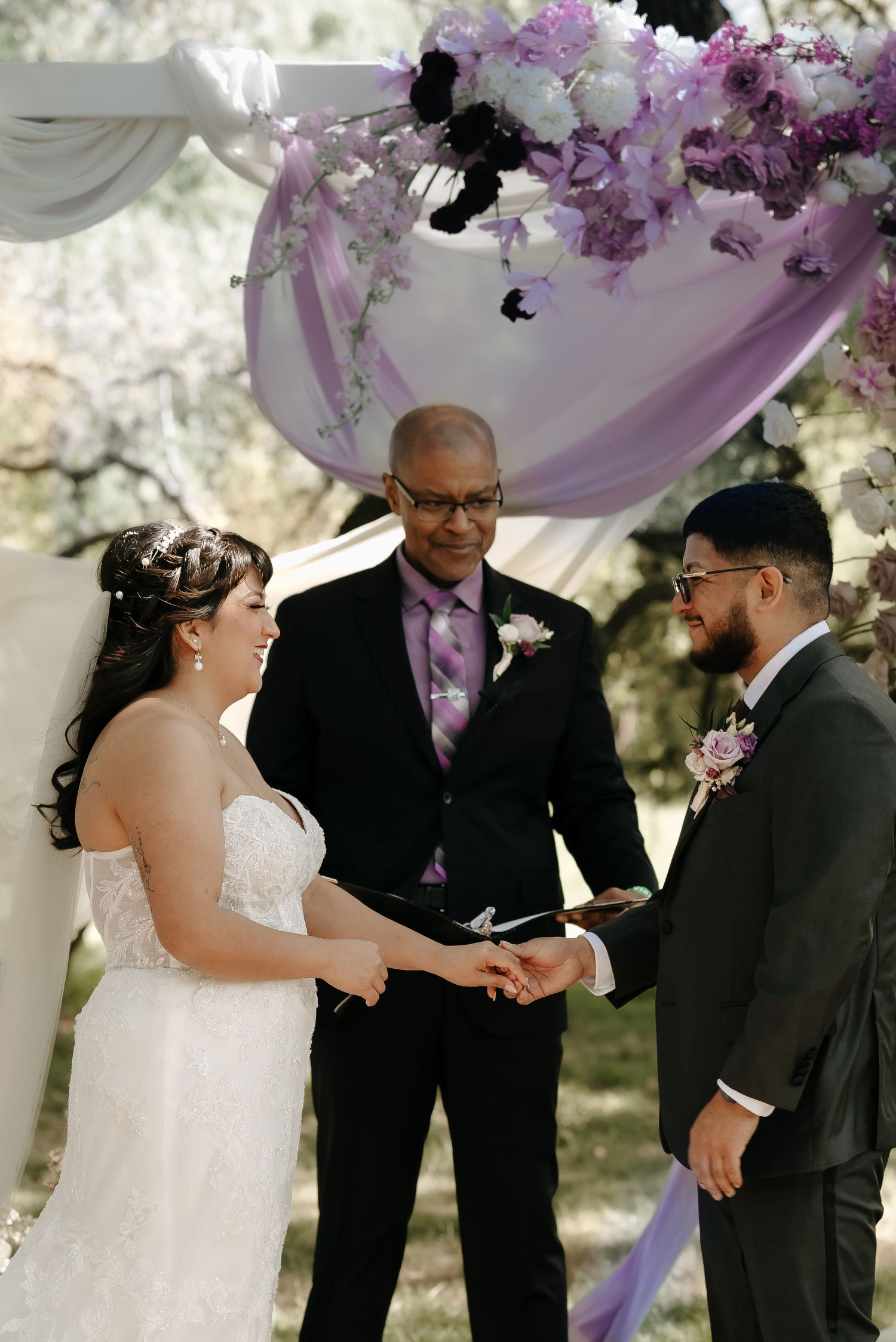 Wedding ceremony with purple and white floral arch decorations in natural setting.