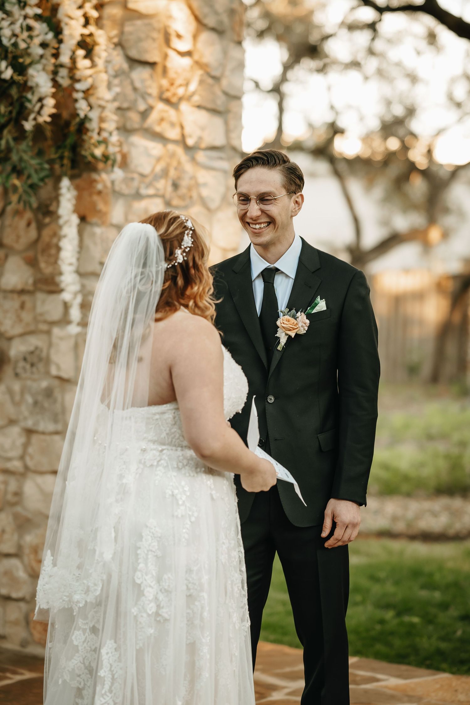 Romantic wedding moment between newlyweds sharing joyful laughter against stone wall and sunset backdrop.