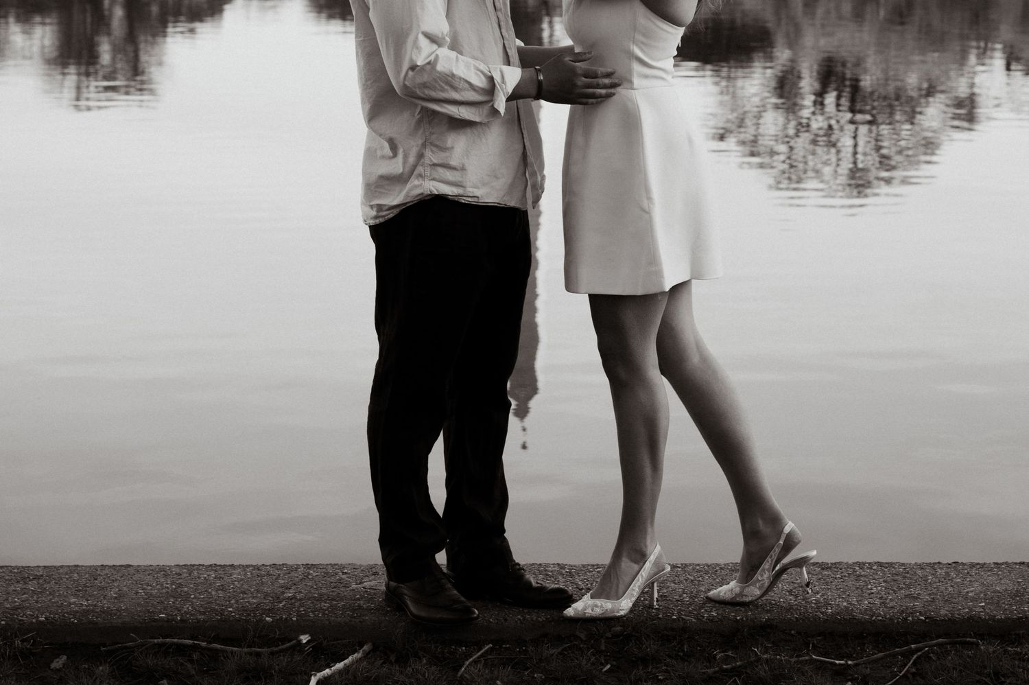 Black and white photo of a couple standing close together by a reflective lake, woman in white dress and heels.