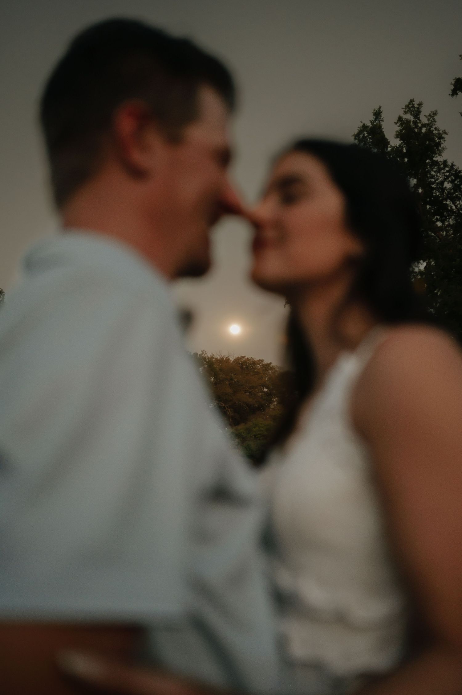 A couple shares an intimate moment at sunset with trees in the background.