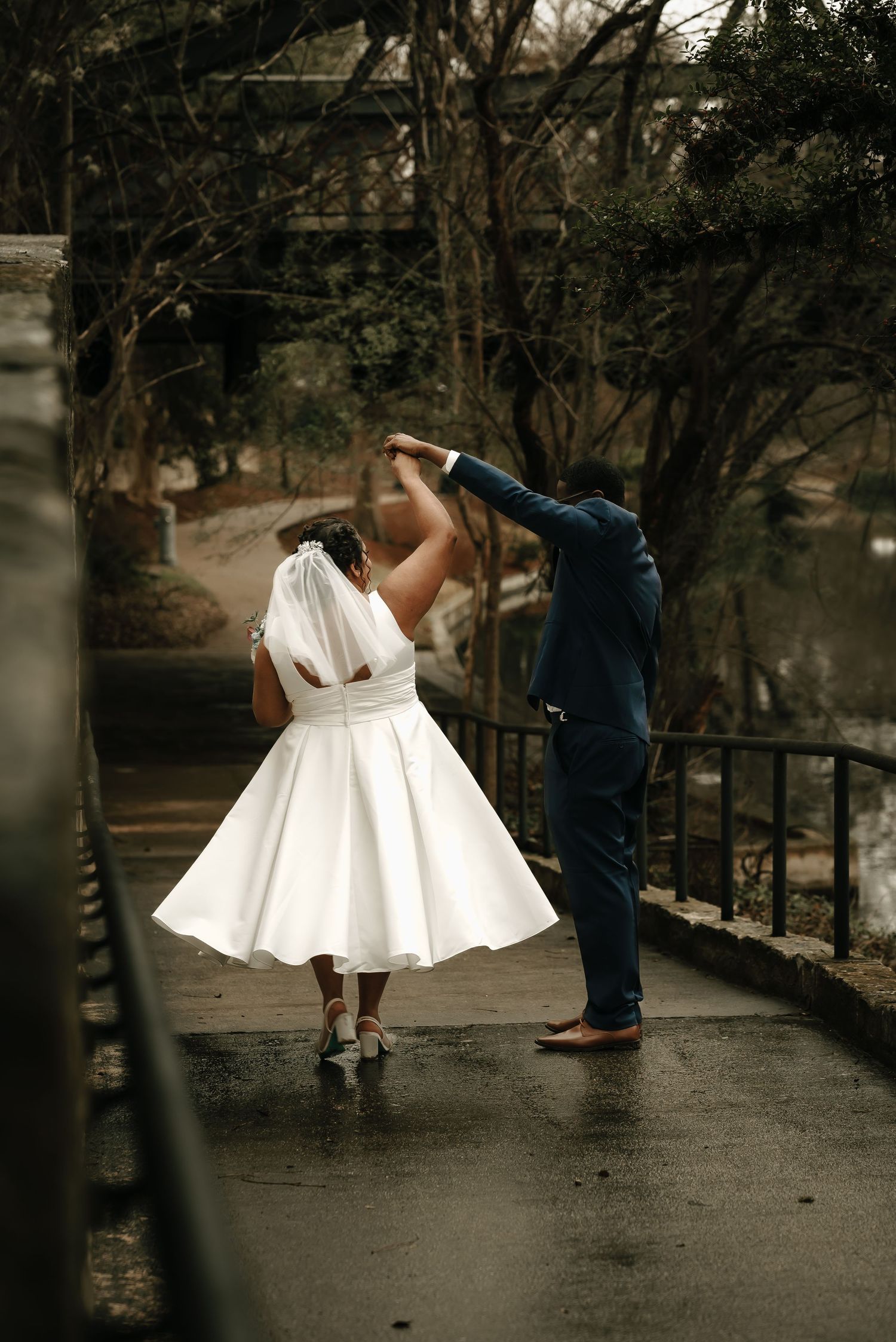 Wedding couple dances playfully on a wet pathway surrounded by bare trees.
