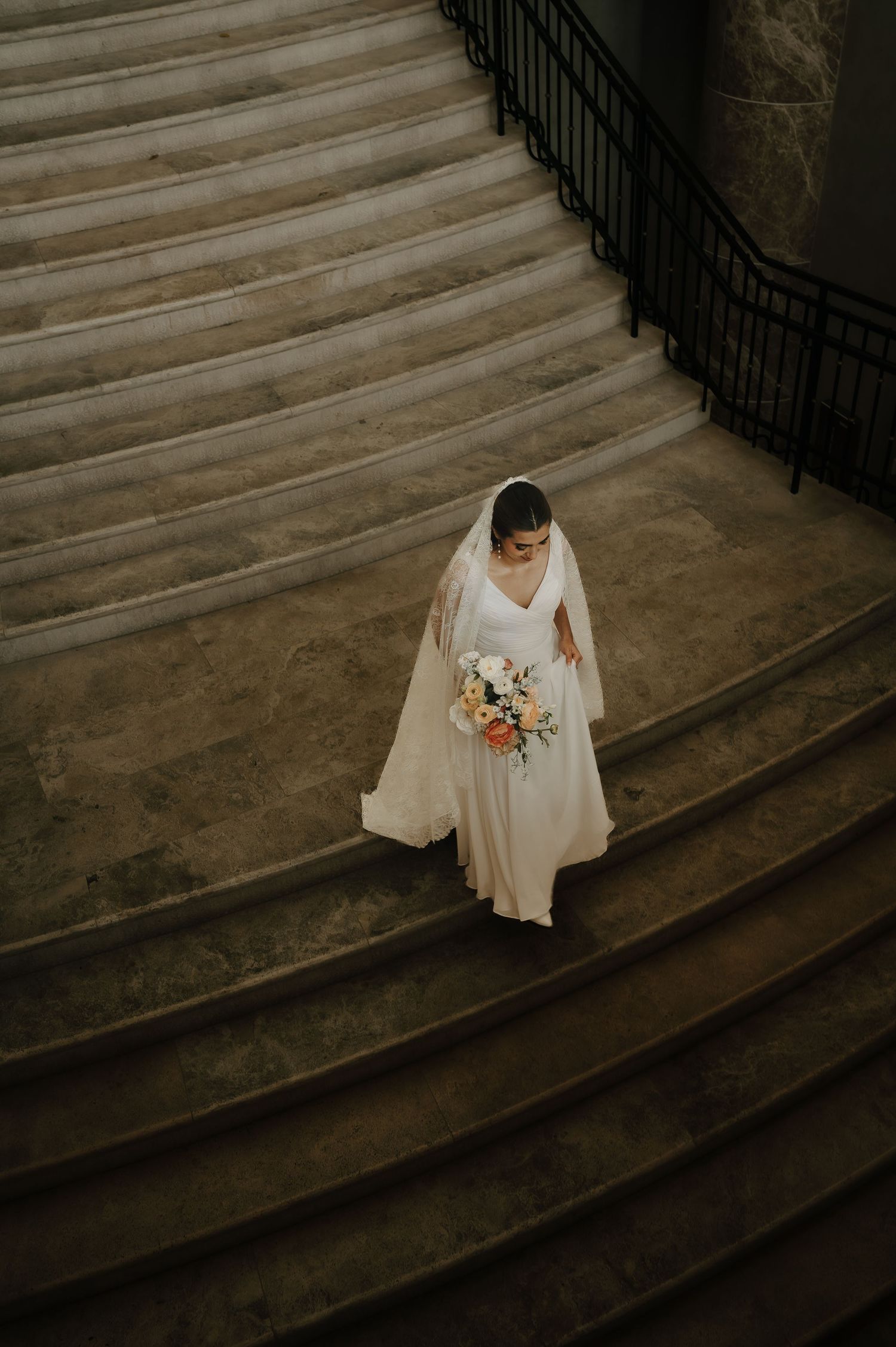A bride in a flowing white dress descending elegant stone steps in dramatic lighting.