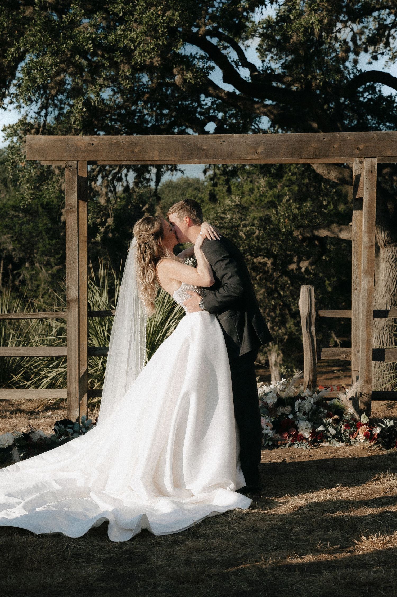Romantic wedding couple embraces under wooden pergola during outdoor ceremony.