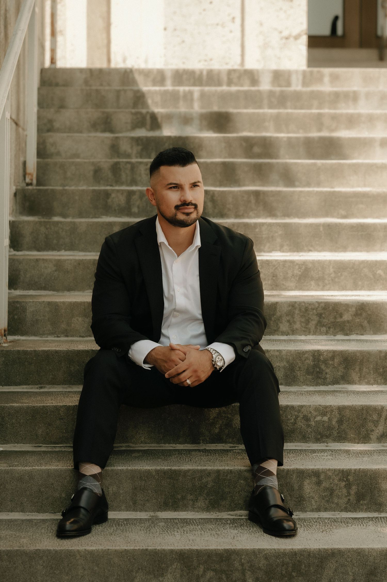 A groom in a black suit sits thoughtfully on concrete steps during a wedding photoshoot.