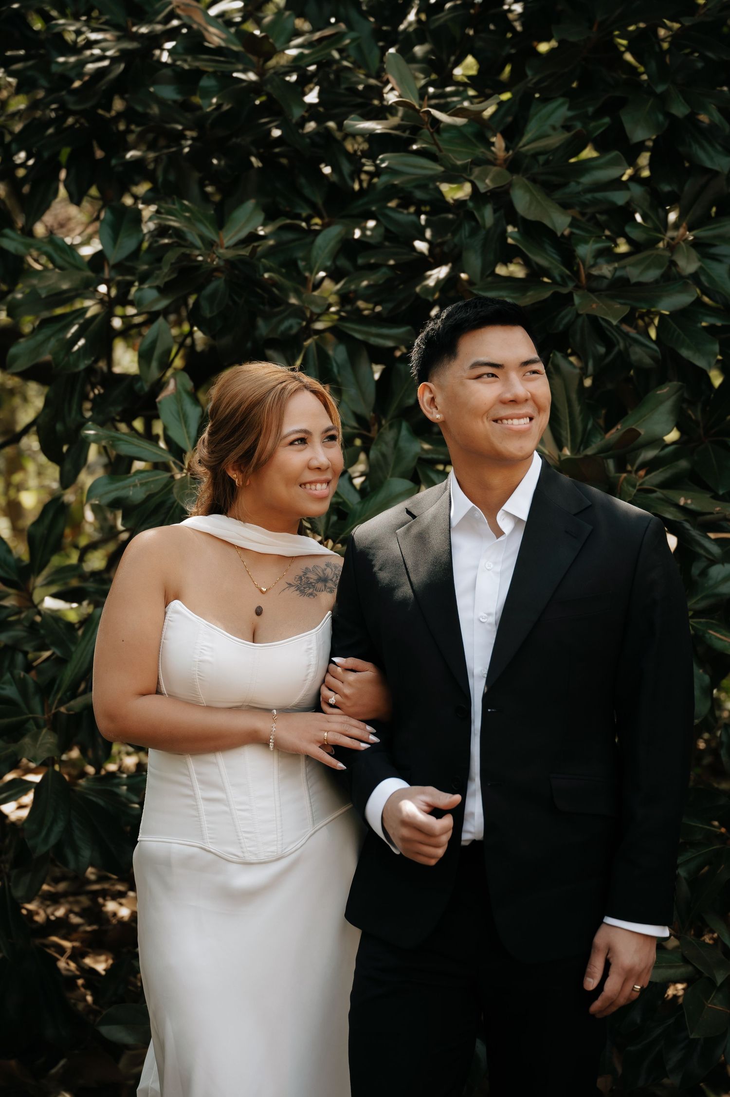 Bride in white gown and groom in black suit smiling together in front of lush green foliage.