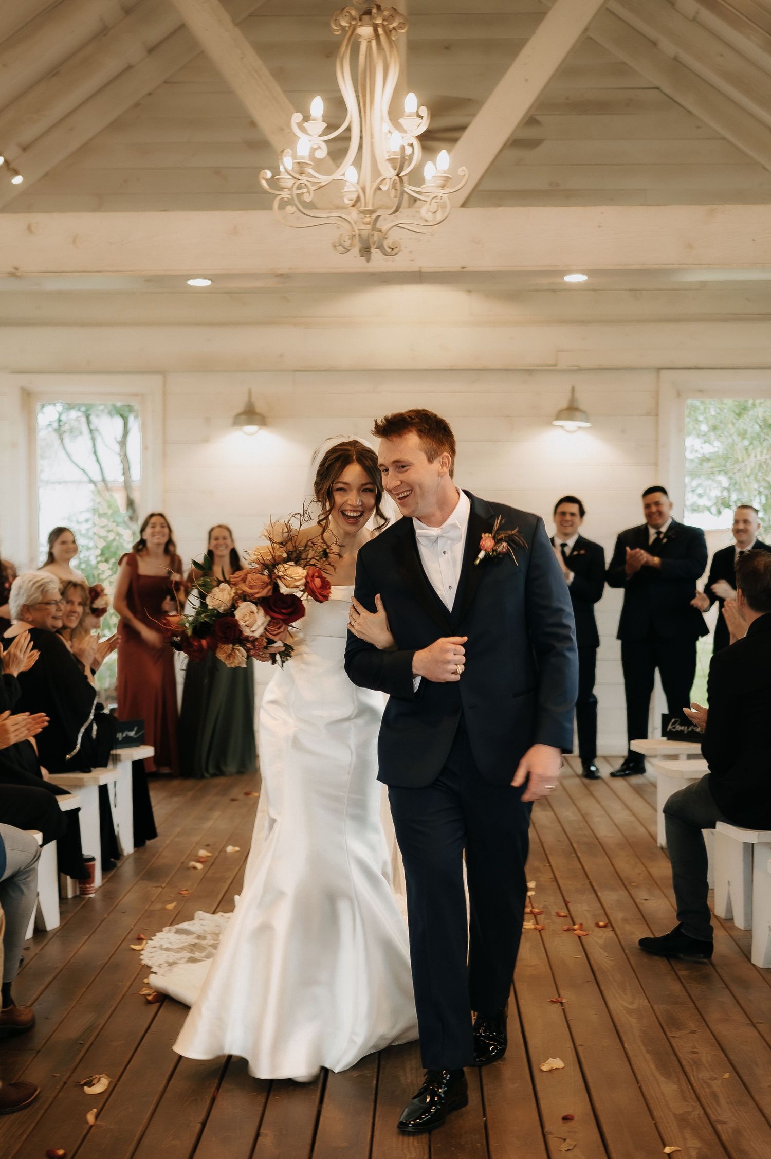 Couple dancing in a rustic white reception hall with wooden floors and elegant chandeliers.