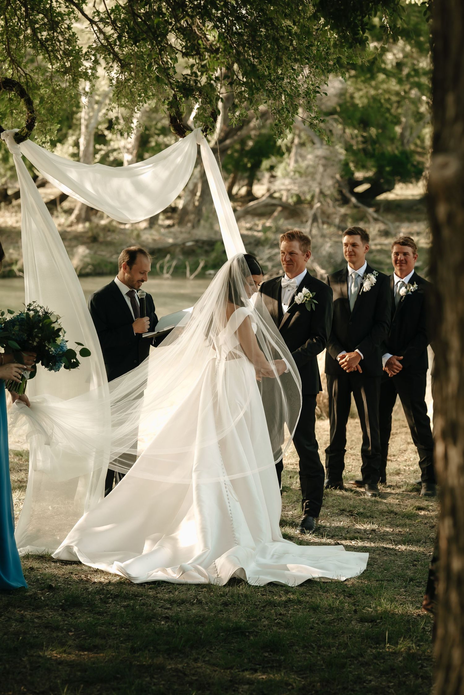 White wedding dresses flowing in the breeze during an outdoor ceremony with guests watching.