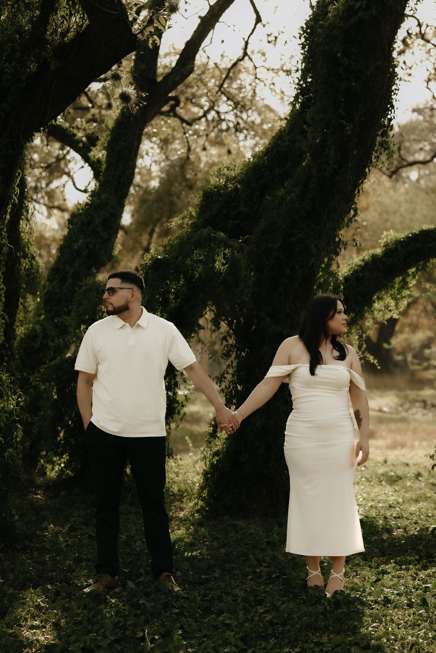 A romantic outdoor photo session showing a couple in white outfits walking among trees in golden light.