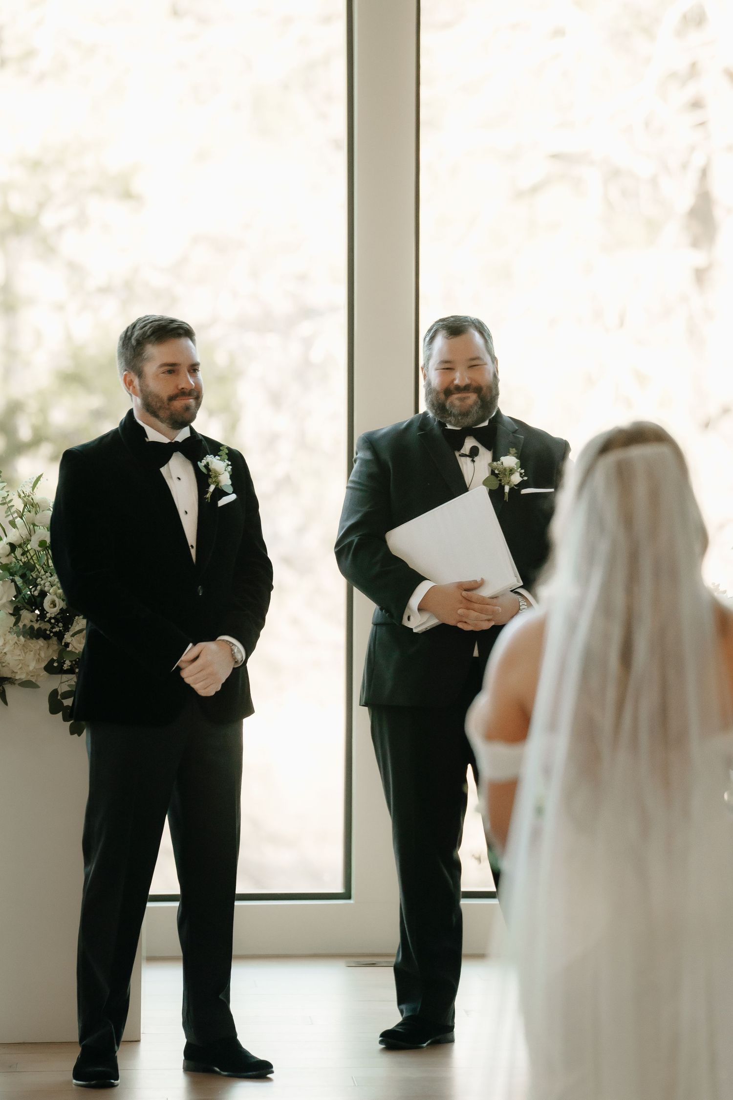 Wedding ceremony participants in formal attire standing in front of bright windows.