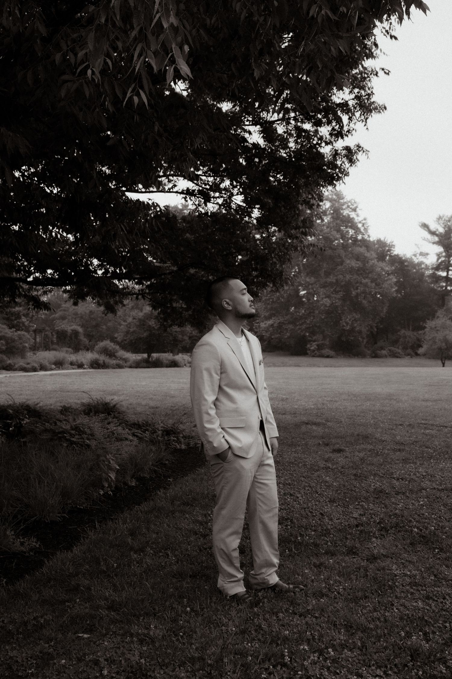 Man in light suit standing under large tree in black and white outdoor portrait photography.