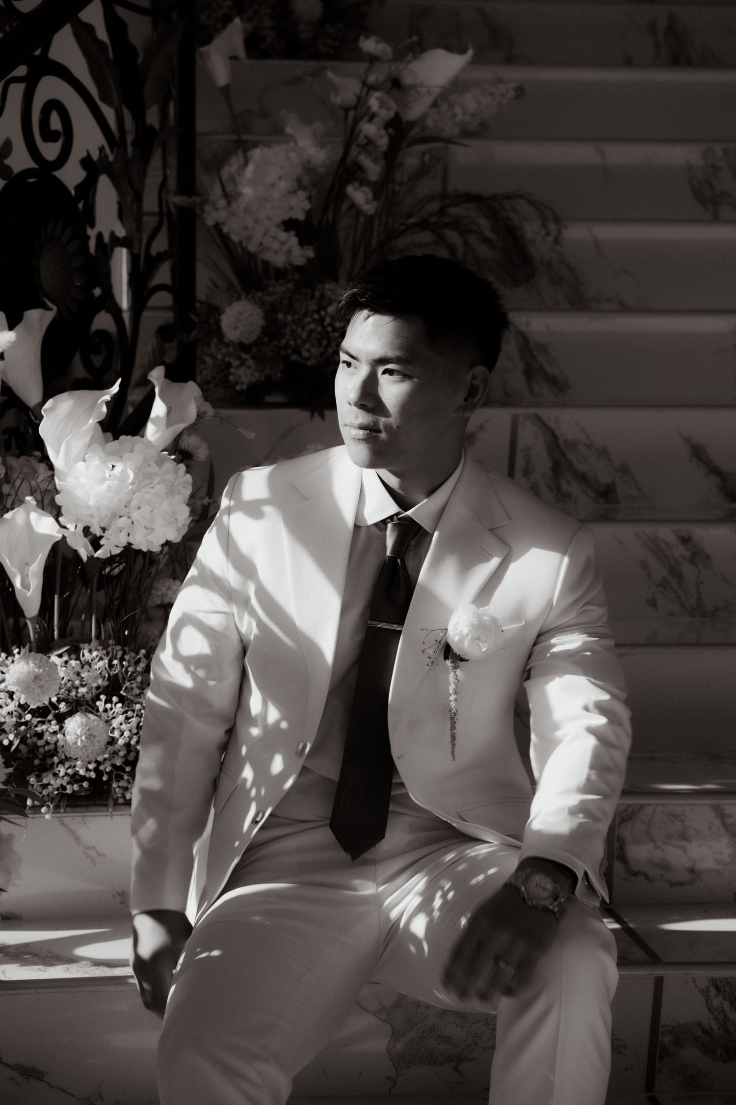 Groom in a white suit and dark tie sits pensively on marble steps in a black and white portrait.