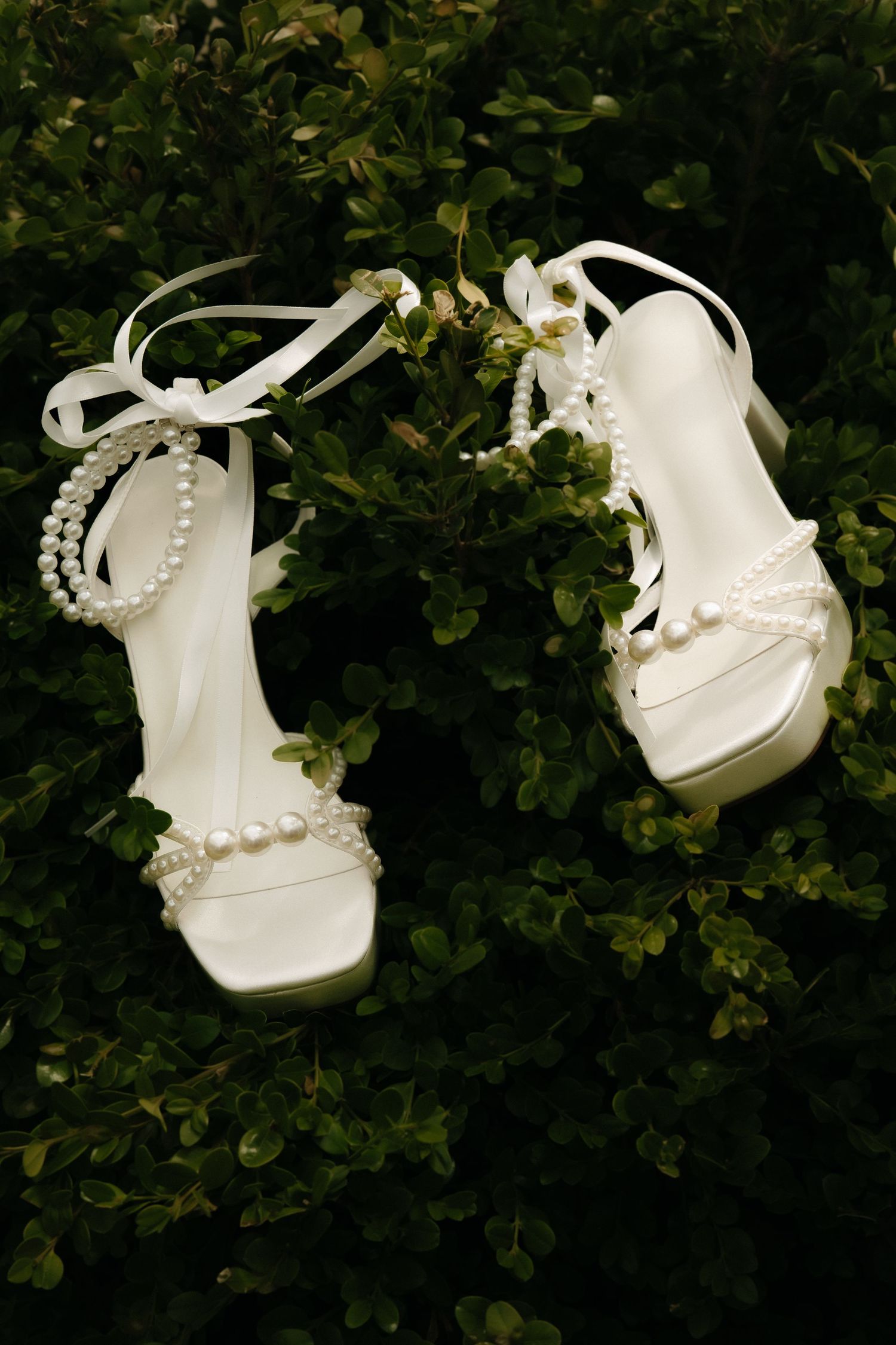 White bridal heeled sandals adorned with pearl embellishments and ribbon ankle ties, displayed on lush green foliage.