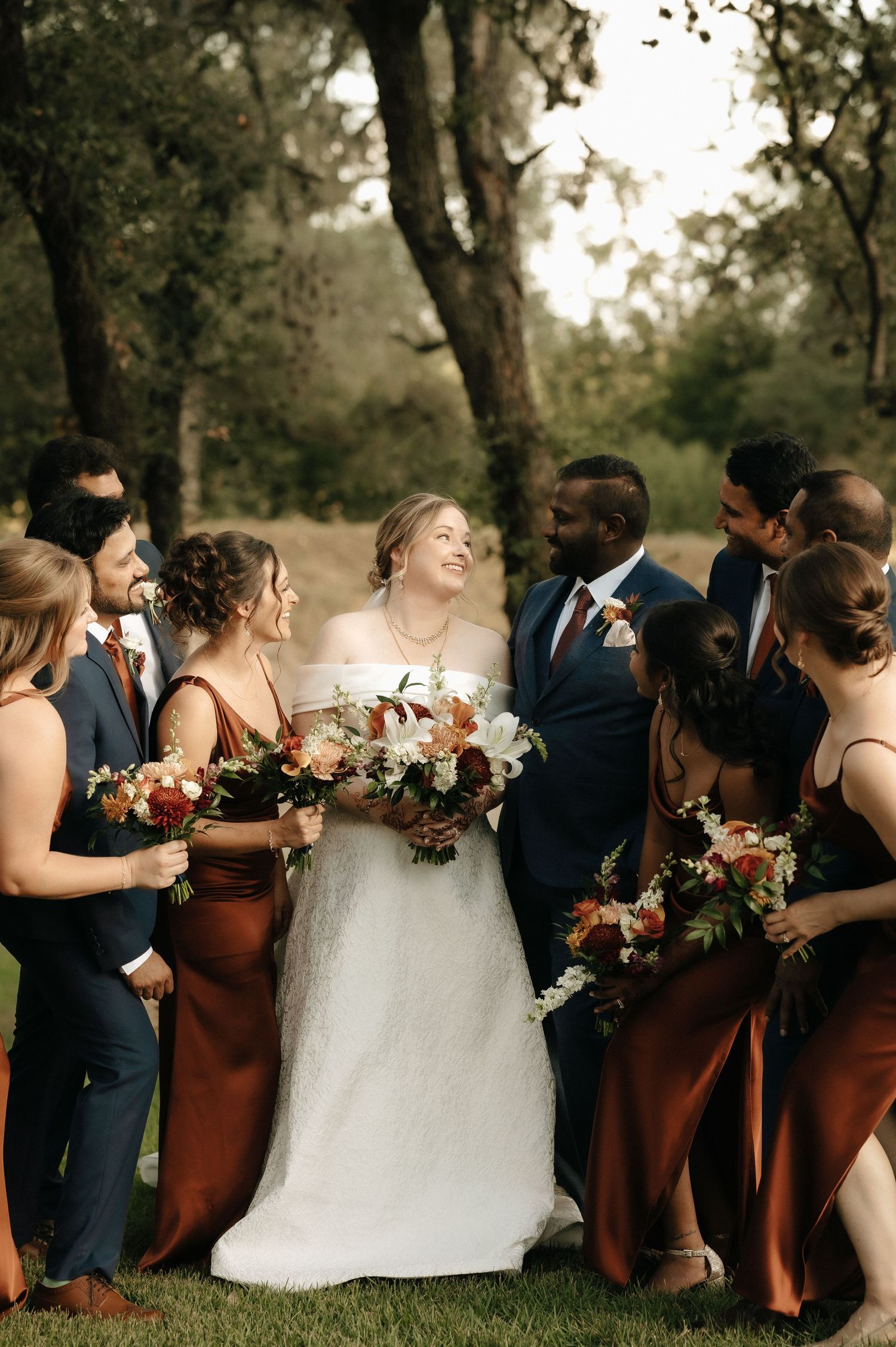 Wedding party in navy blue suits and rust-colored dresses poses together outdoors holding floral bouquets.