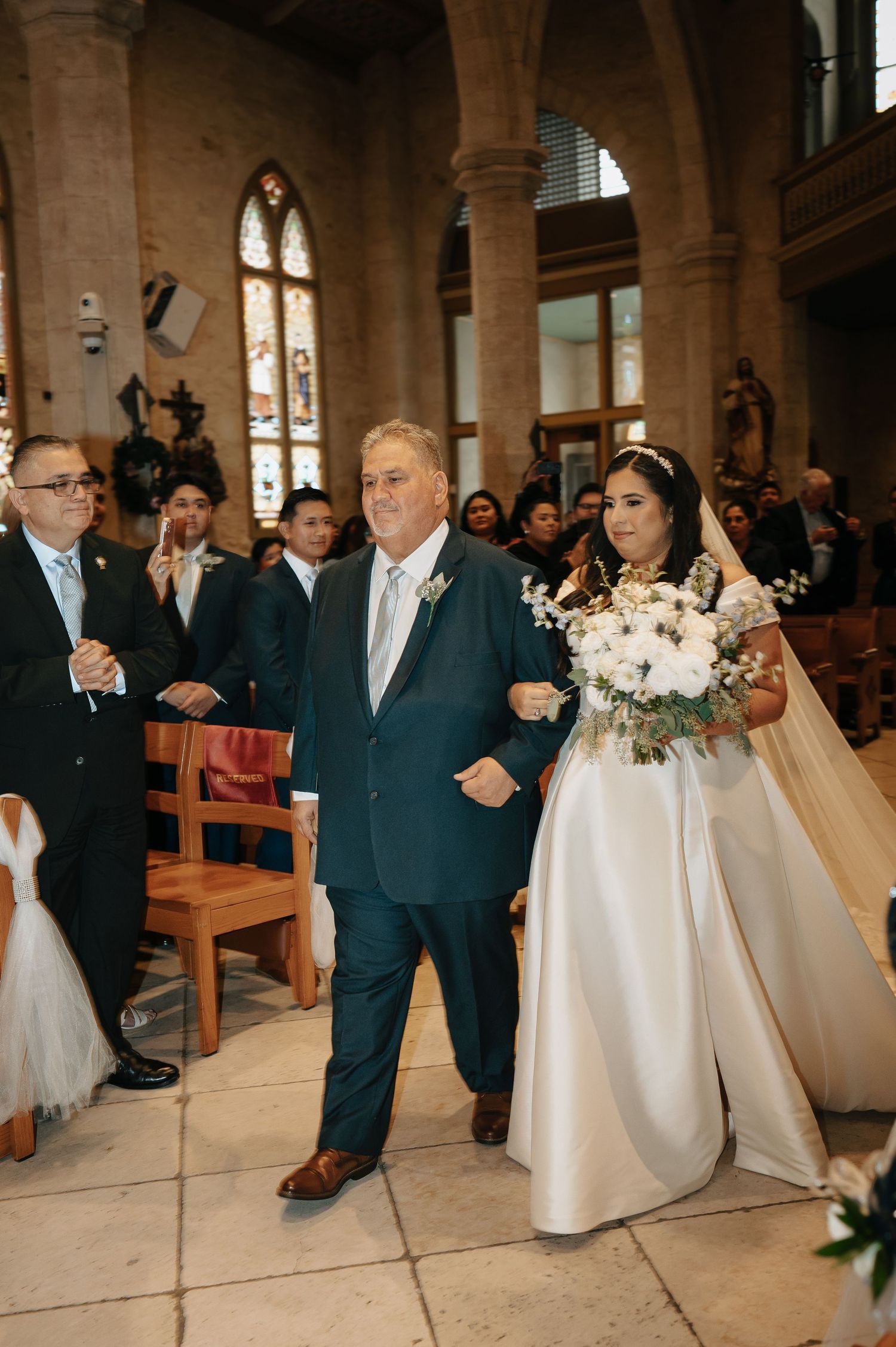 Wedding ceremony procession in a traditional church with guests in navy suits lining the aisle.