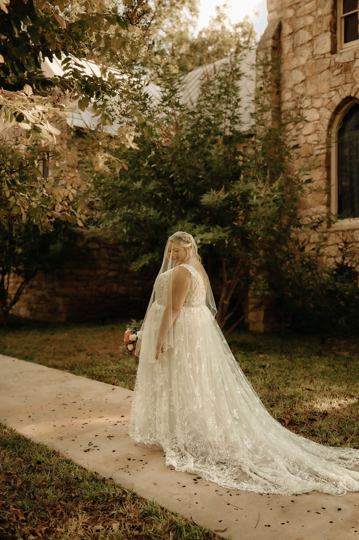 A flowing lace wedding dress and veil trail elegantly along a stone path in an outdoor garden setting.
