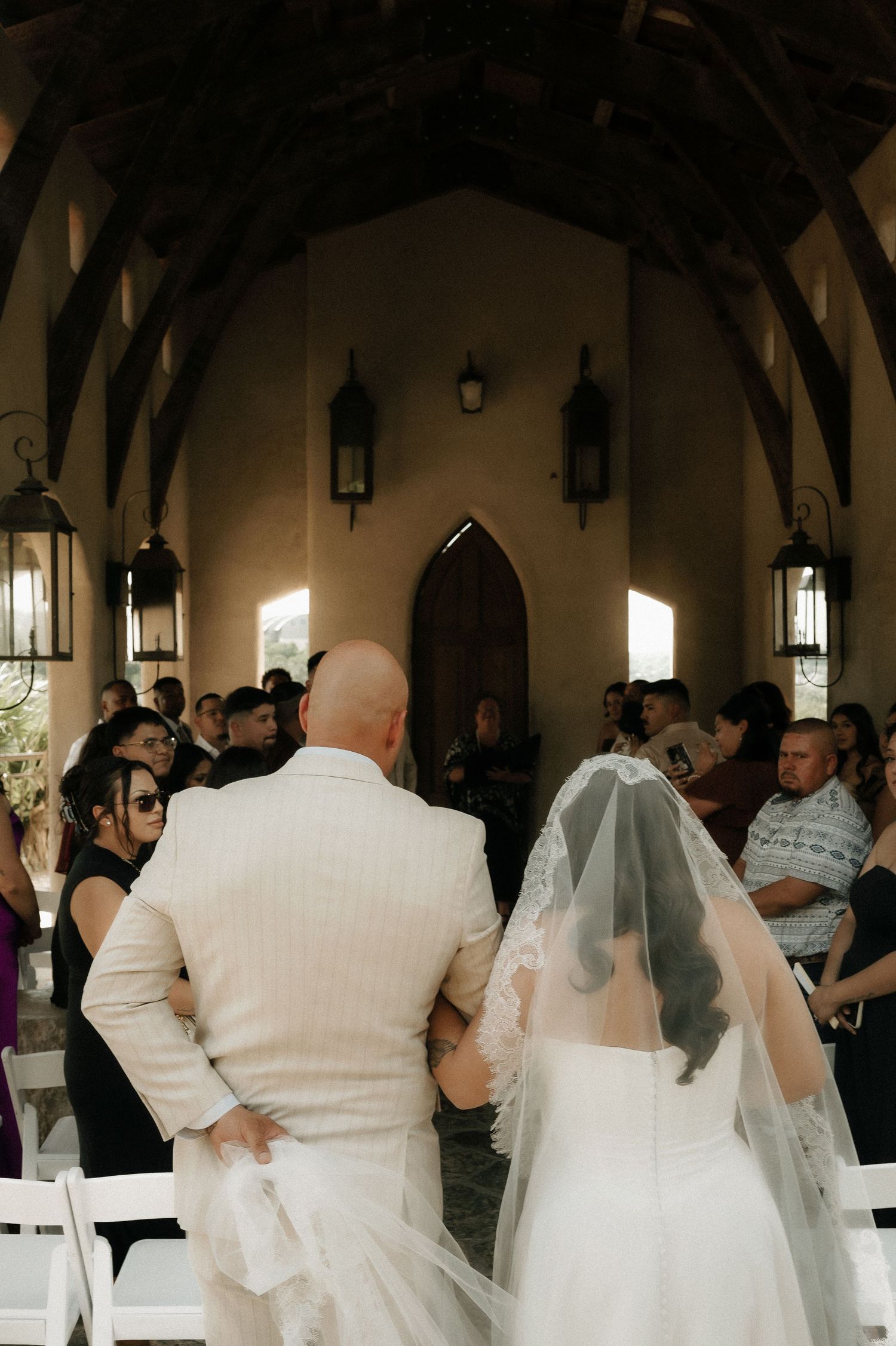 Wedding guests gather inside a rustic chapel with hanging lanterns and arched doorways.