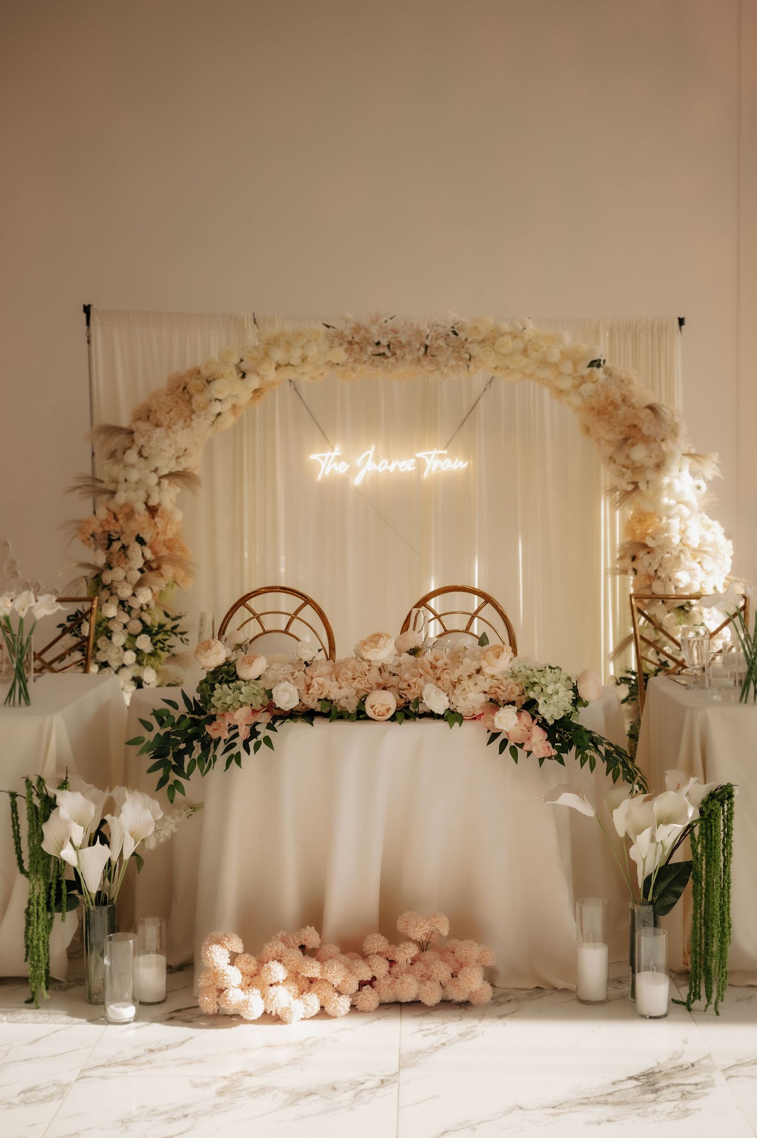 Elegant wedding sweetheart table with floral arch, neon sign, blush roses, white draping, and candles on marble floor.