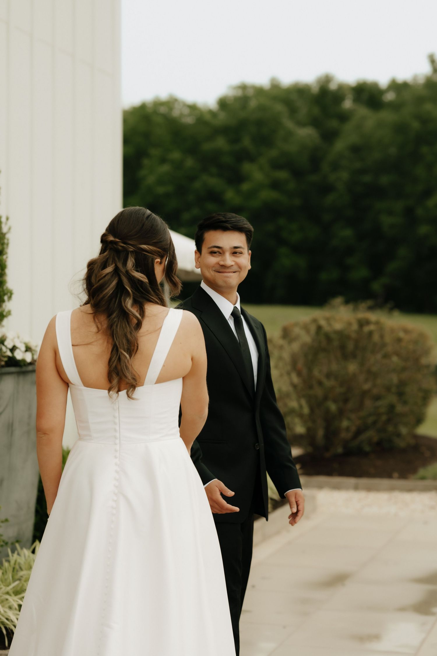 Groom in black suit smiles at bride in white wedding dress during first look at outdoor venue.