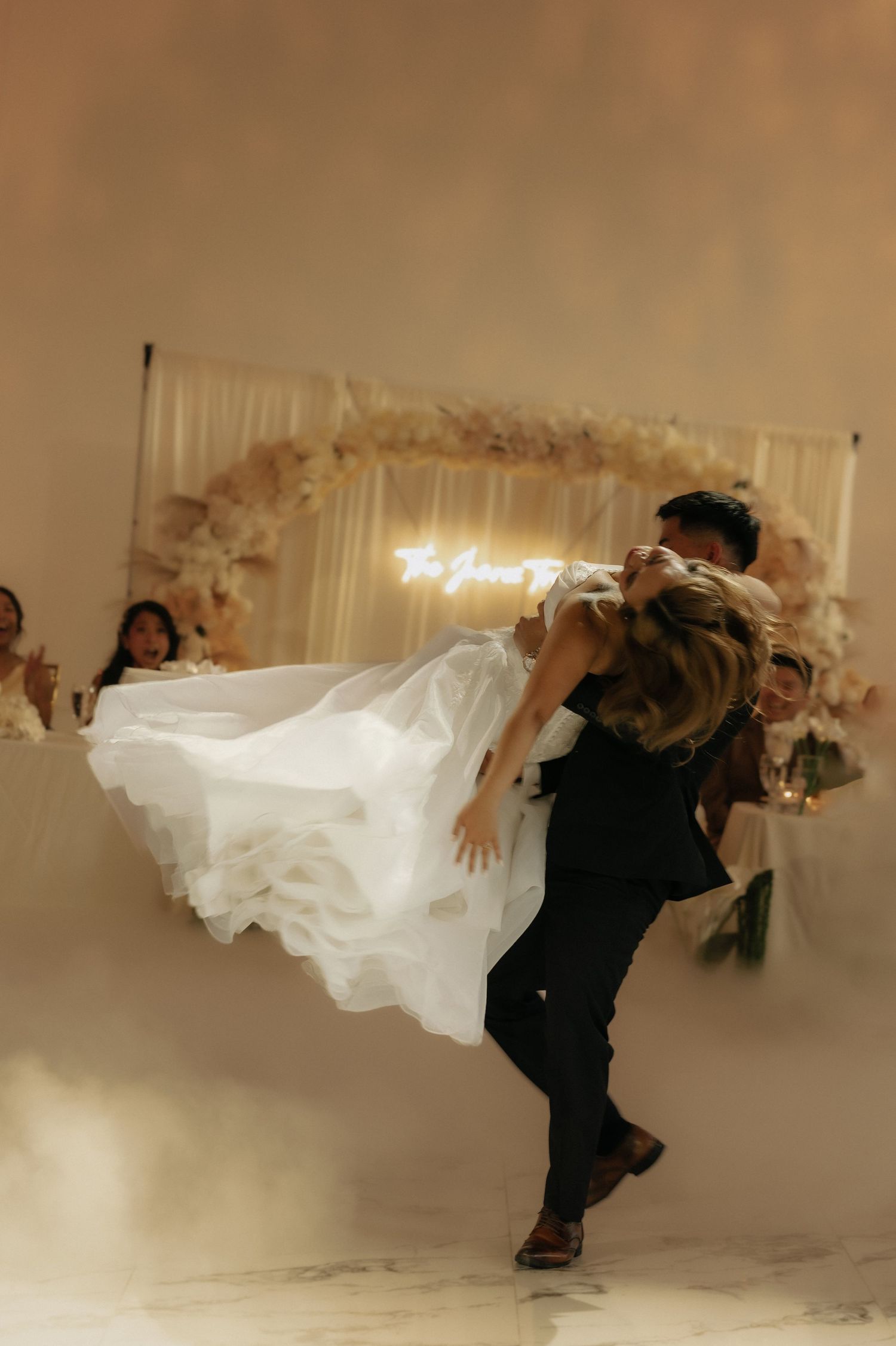 Groom lifting bride in wedding dress during first dance with floral arch backdrop and soft fog on dance floor.
