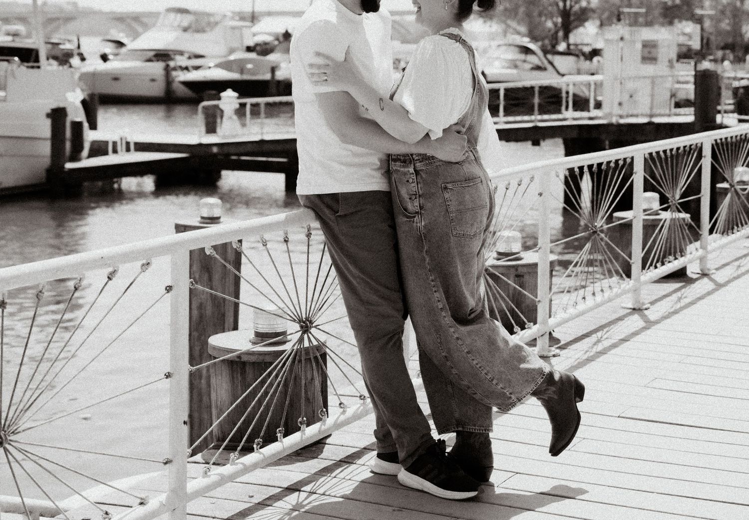 Couple embracing romantically on a wooden marina dock with boats in the background, black and white photo.