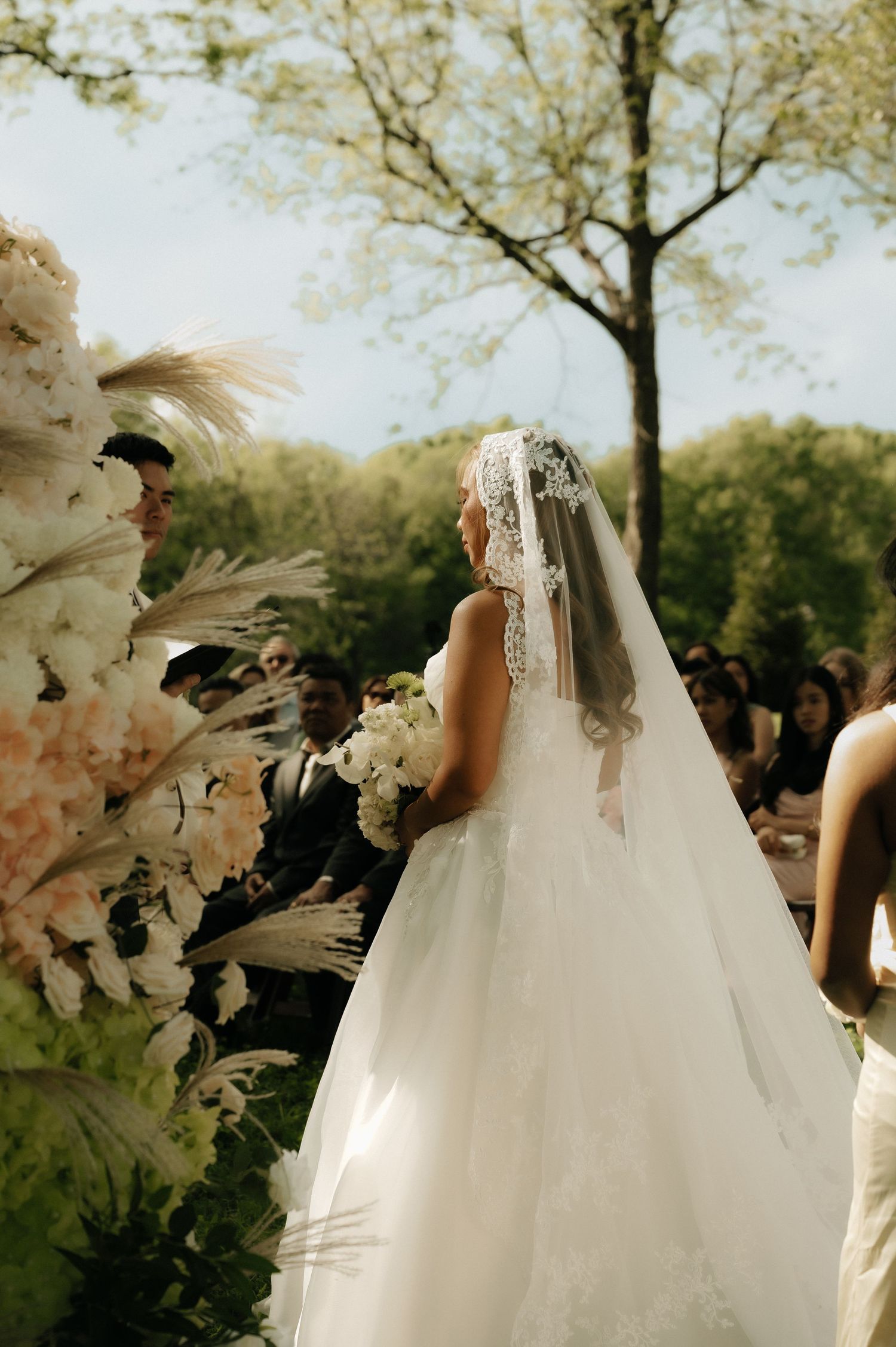 Bride in white ball gown and lace veil walks down outdoor aisle with floral arrangements at garden wedding ceremony.