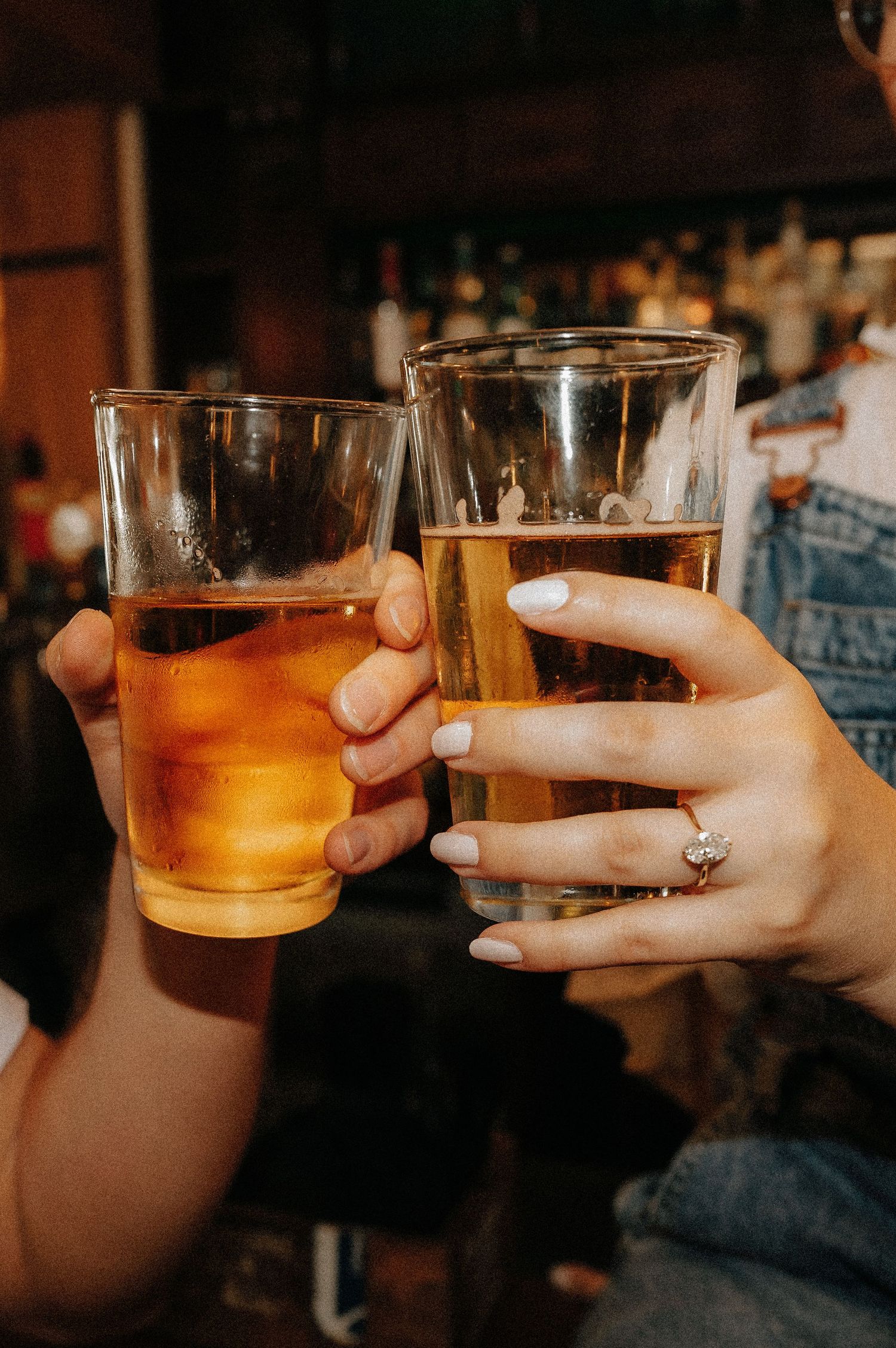 Two people toasting with glasses of beer at a bar, one wearing a diamond engagement ring.