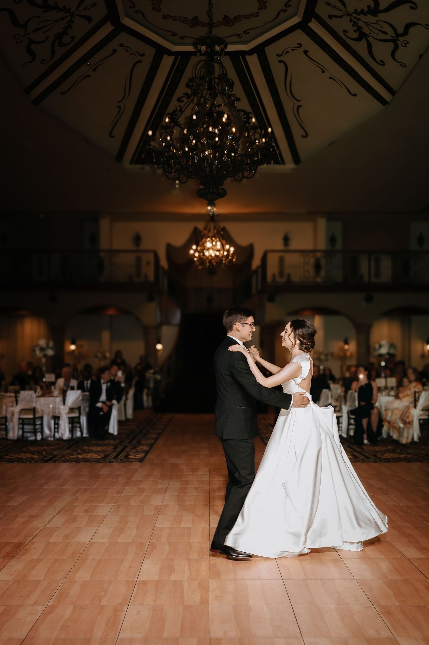 A couple shares a romantic dance on a wooden ballroom floor under elegant chandeliers at their wedding reception.