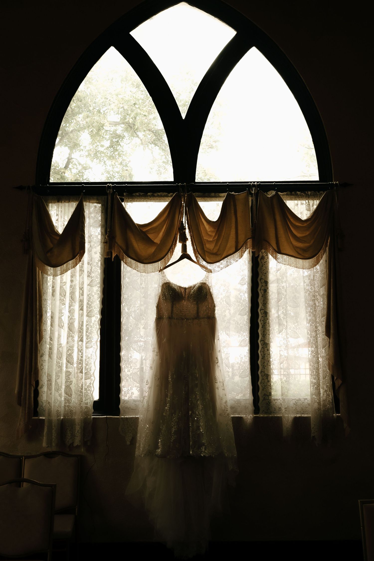 Silhouette of wedding dress hanging in arched window with dramatic lighting.