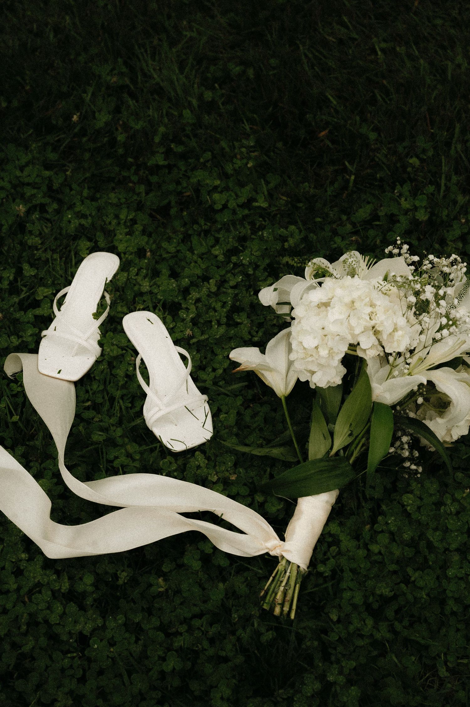 White bridal heels with ribbon straps beside a white floral bouquet with greenery on lush green clover grass.