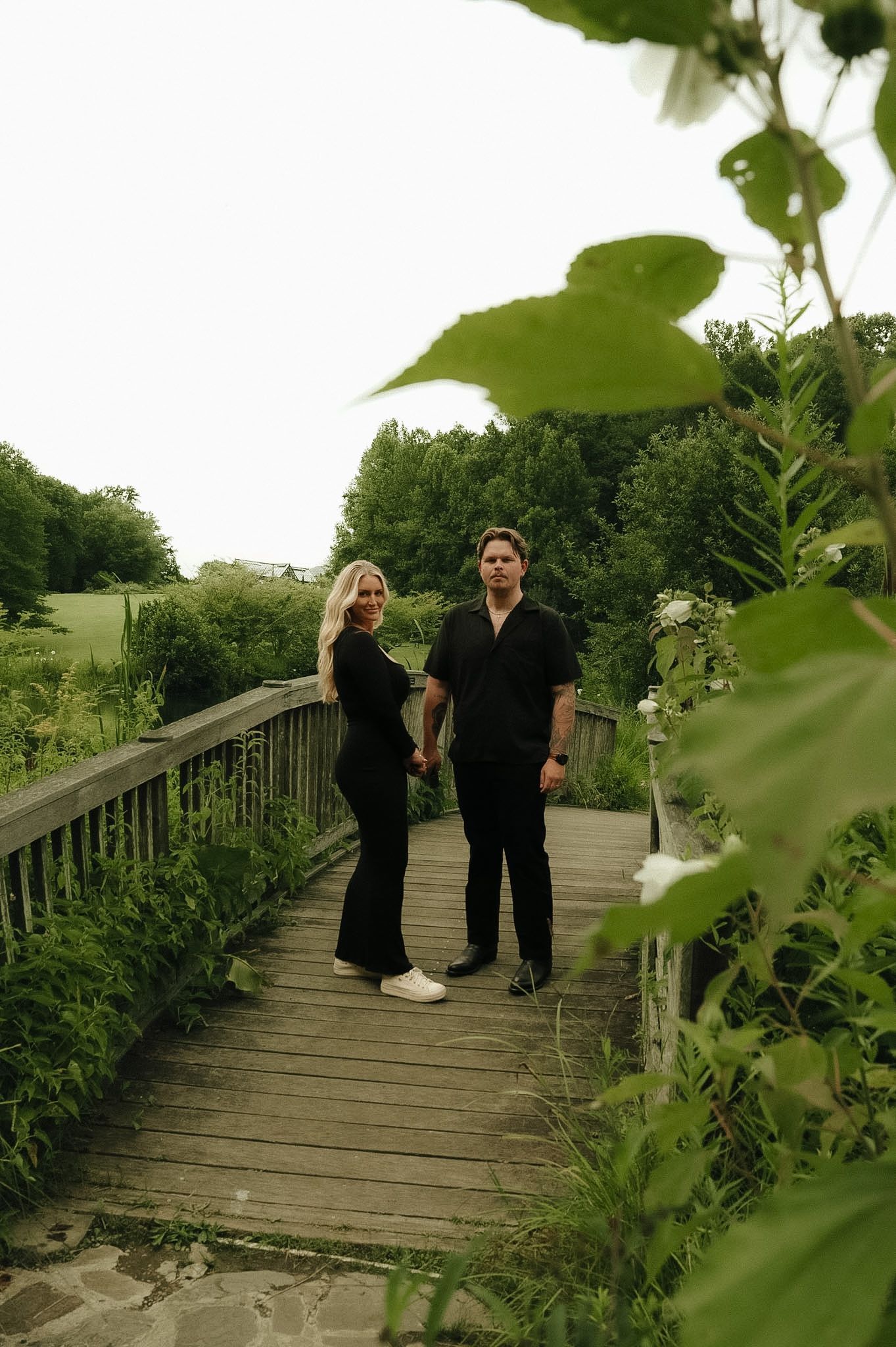 Couple in black outfits holding hands on a wooden bridge surrounded by lush green garden foliage.