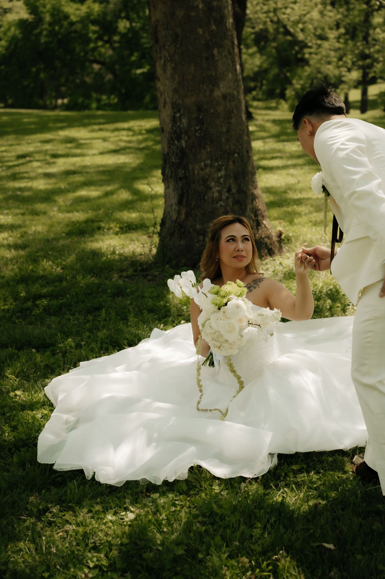 Bride in white gown sits on green grass leaning against tree as groom in white suit reaches for her hand.