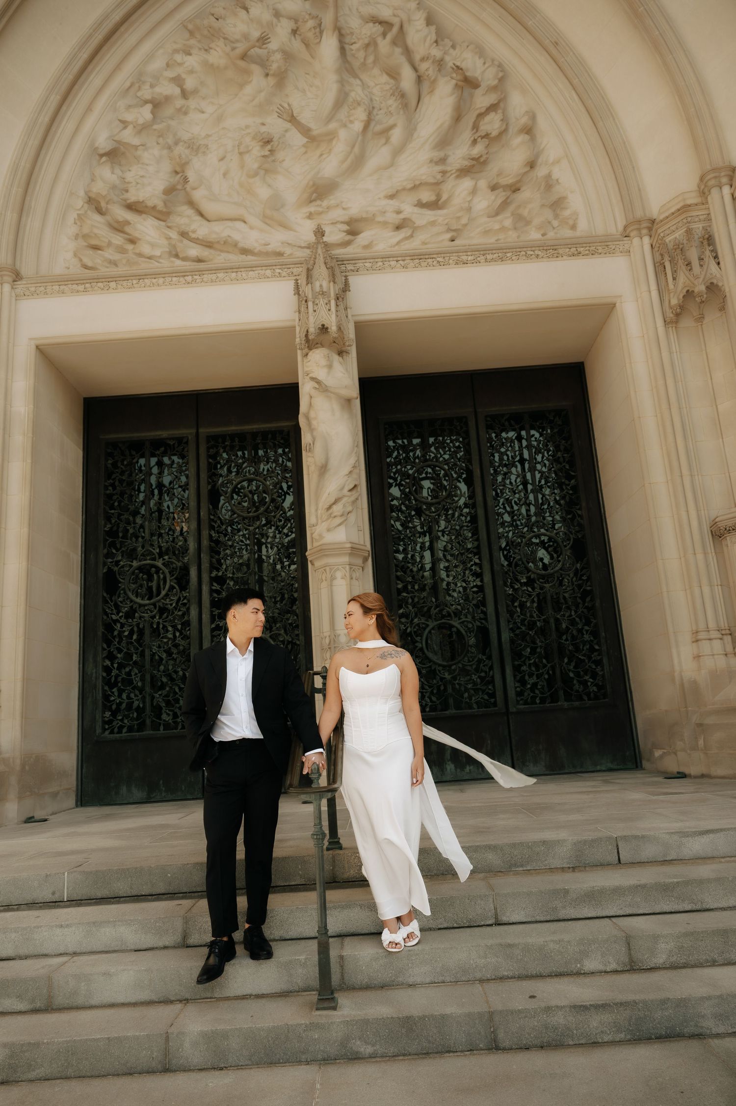 Newlywed couple holding hands on ornate church steps, bride in white gown, groom in black suit.