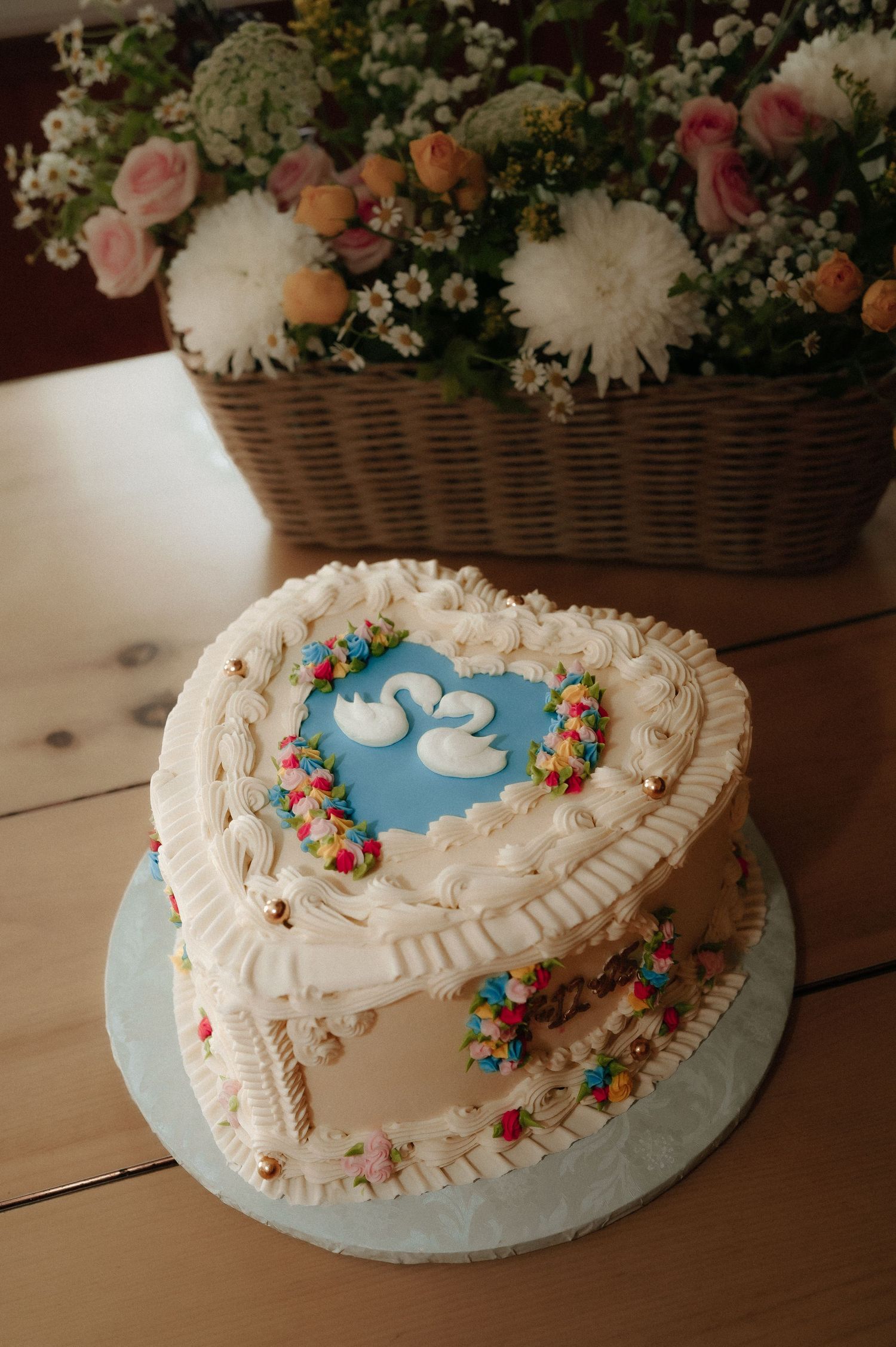 A heart-shaped wedding cake with white frosting and blue details sits beside a floral arrangement.